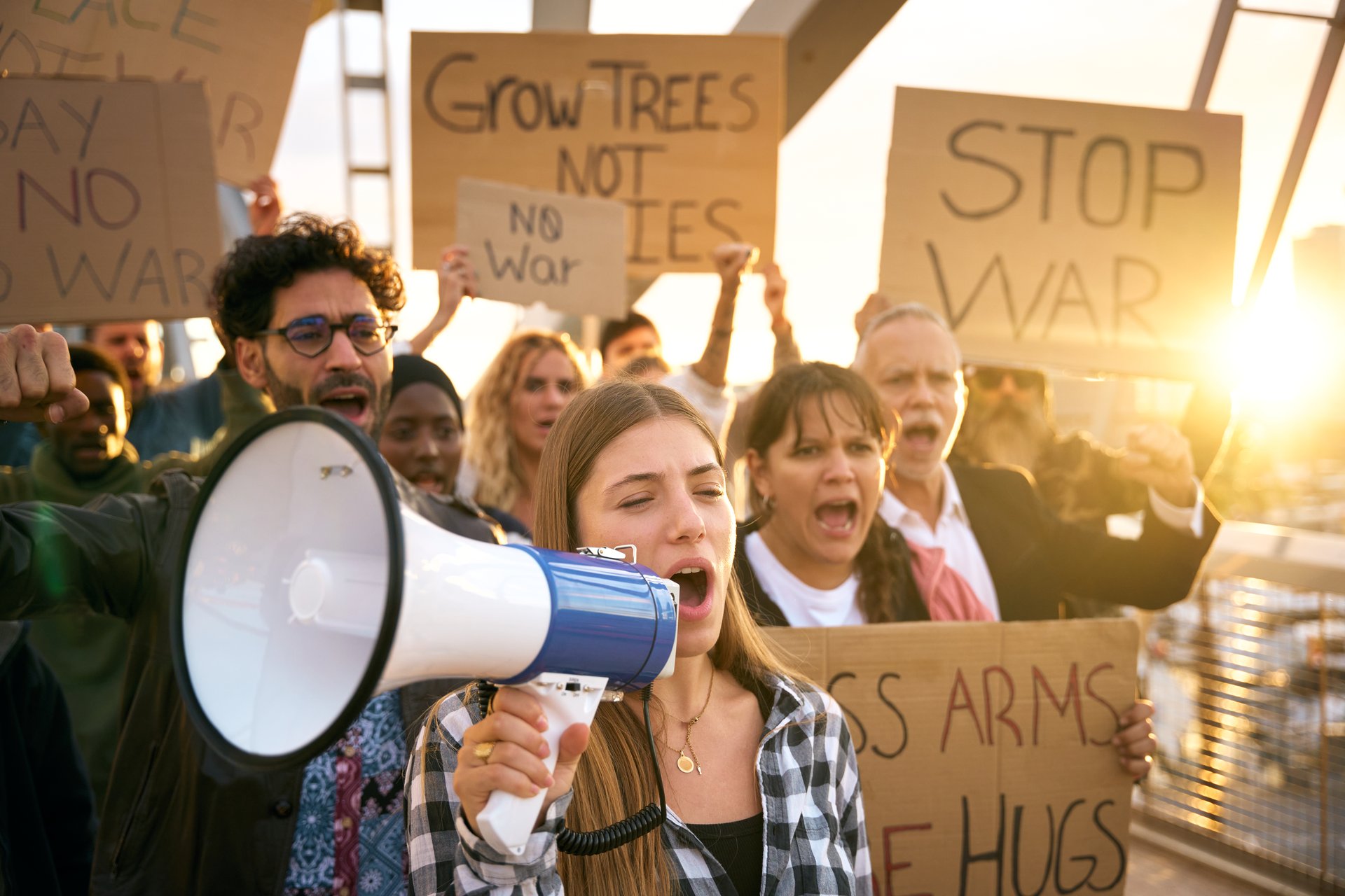 Blonde woman with megaphone in protest with diverse group activist people demonstrate together shouting slogans against wars in world holding banners for peace, love, hugs and not armies