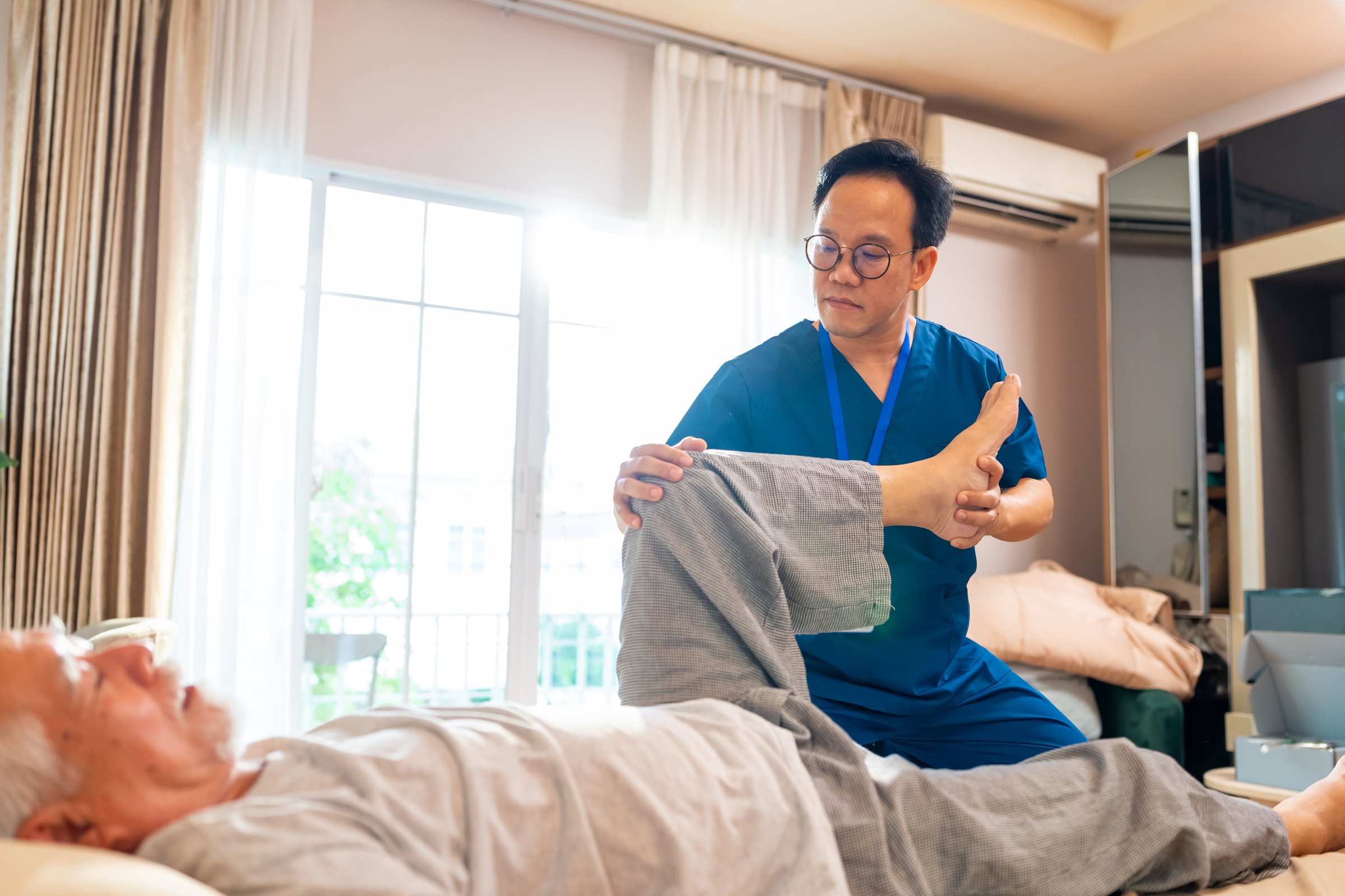 Asian senior man patient doing rehab physical therapy stretching muscle leg and knee with physiotherapist caregiver on bed in bedroom. Home medical caregiving services and elderly people healthcare.