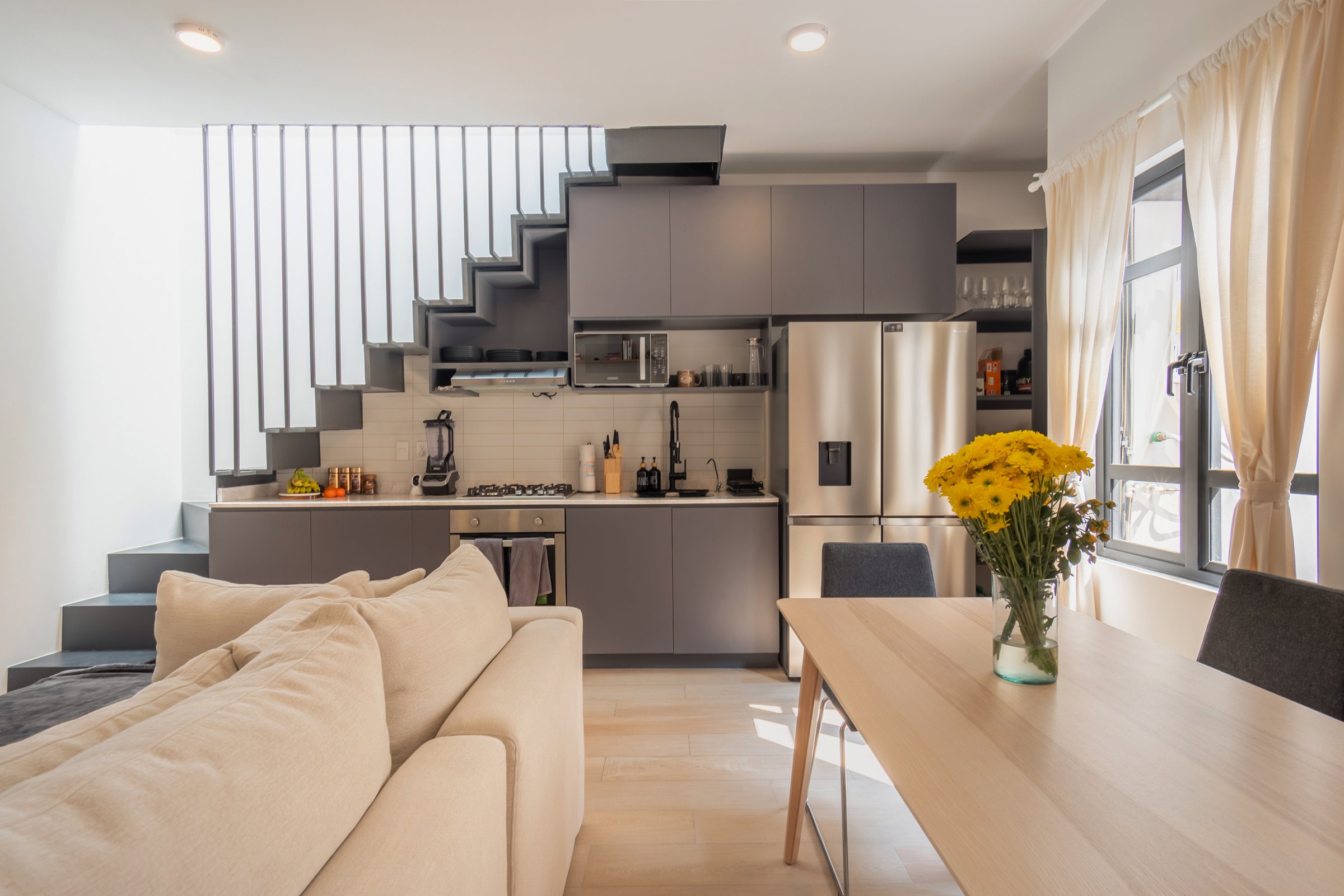 Modern kitchen-living area with gray cabinets, staircase, dining table with flowers, and beige sofa. Natural light through windows.
