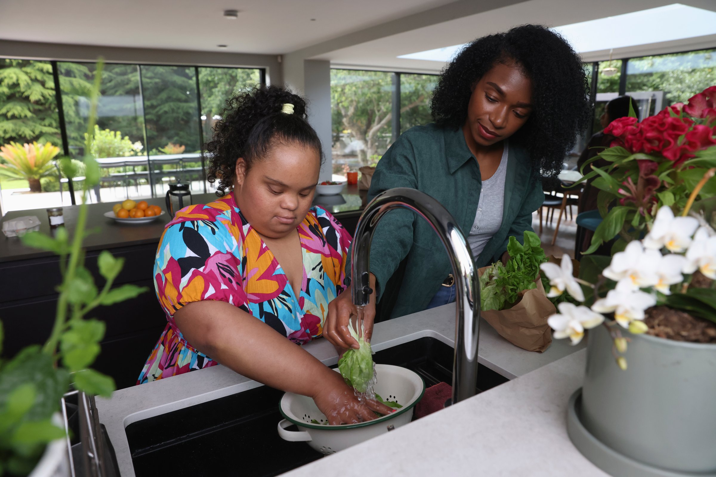 Woman helping young woman with Down syndrome wash greens in kitchen sink
