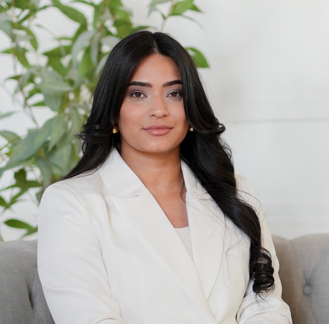 Woman with long dark hair wearing a white blazer, sitting on a gray sofa with plants in the background.