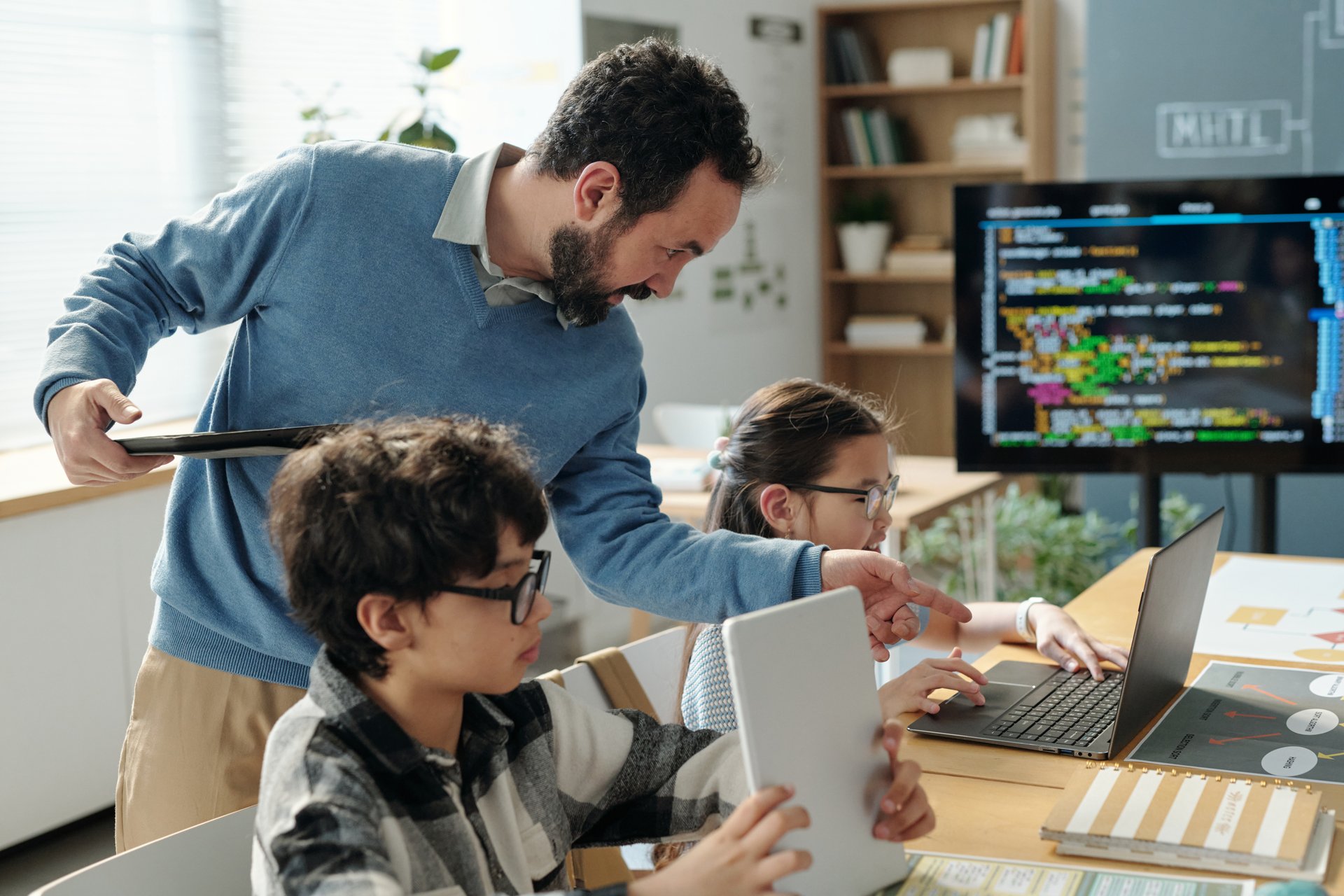Teacher assisting students in learning programming in brightly lit classroom with open laptops and educational tools contributing to interactive atmosphere
