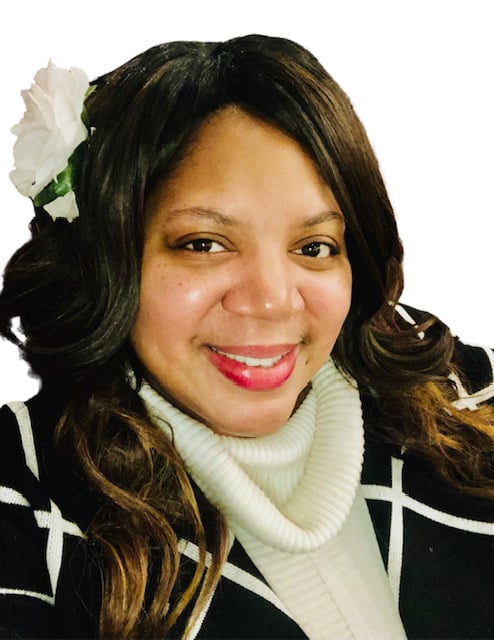 Smiling woman with long hair, wearing a black and white outfit, white flower in hair, against a plain background.