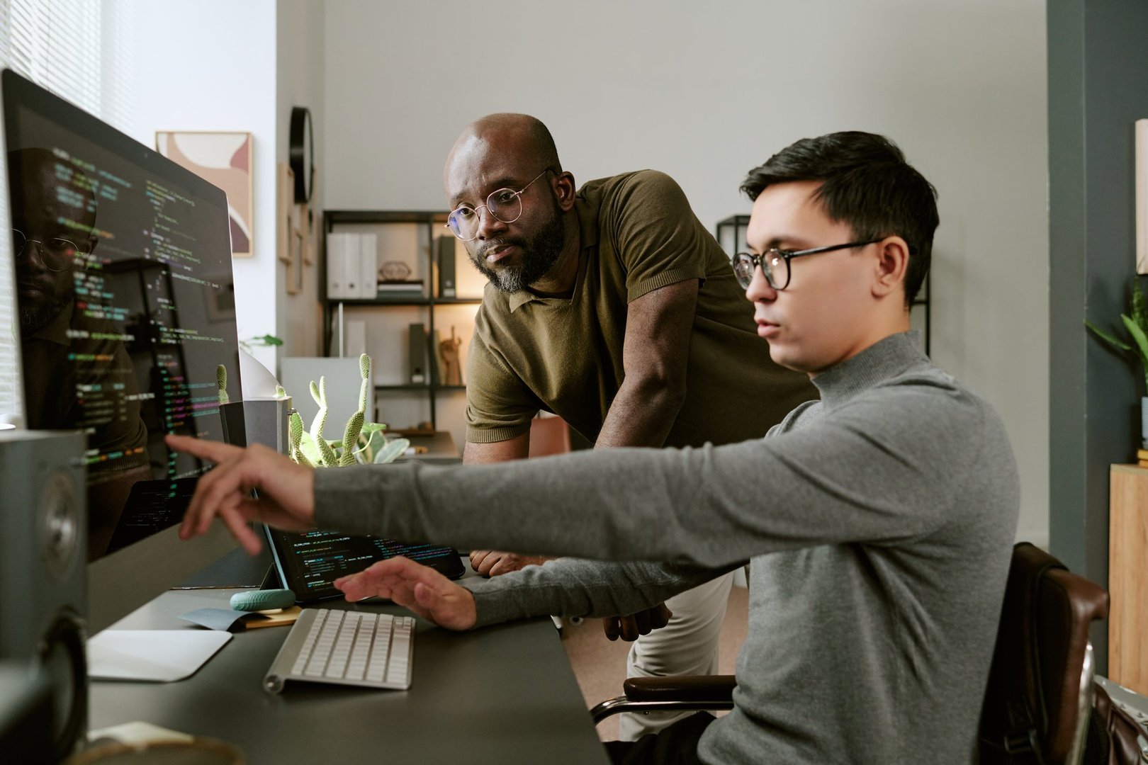 Caucasian young adult man sitting at desk coding on computer while Caucasian young man standing beside him observing screen, both collaborating on software development project in modern office