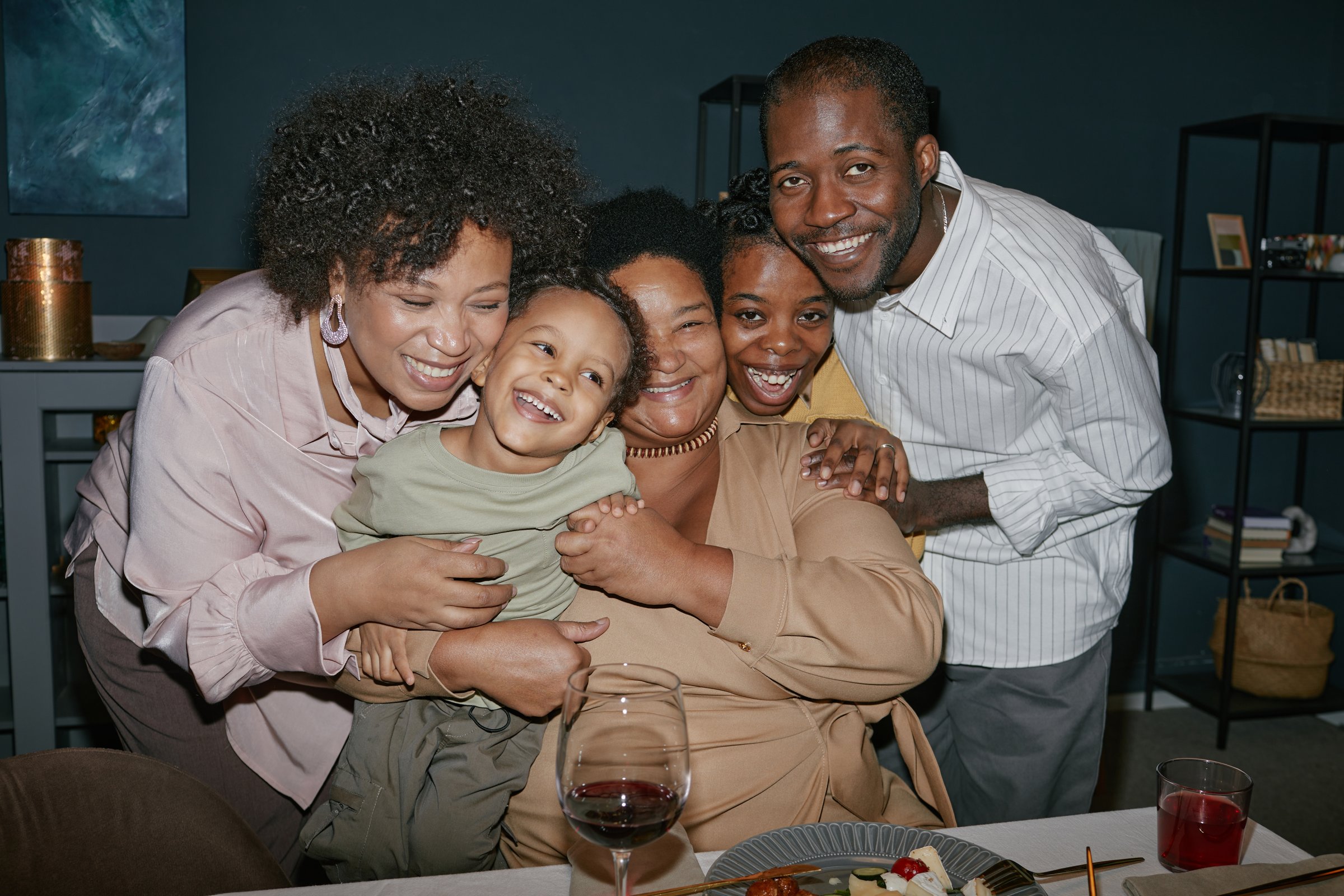 Portrait of smiling Black family members embracing and enjoying quality time at dining table during festive gathering at home, camera flash
