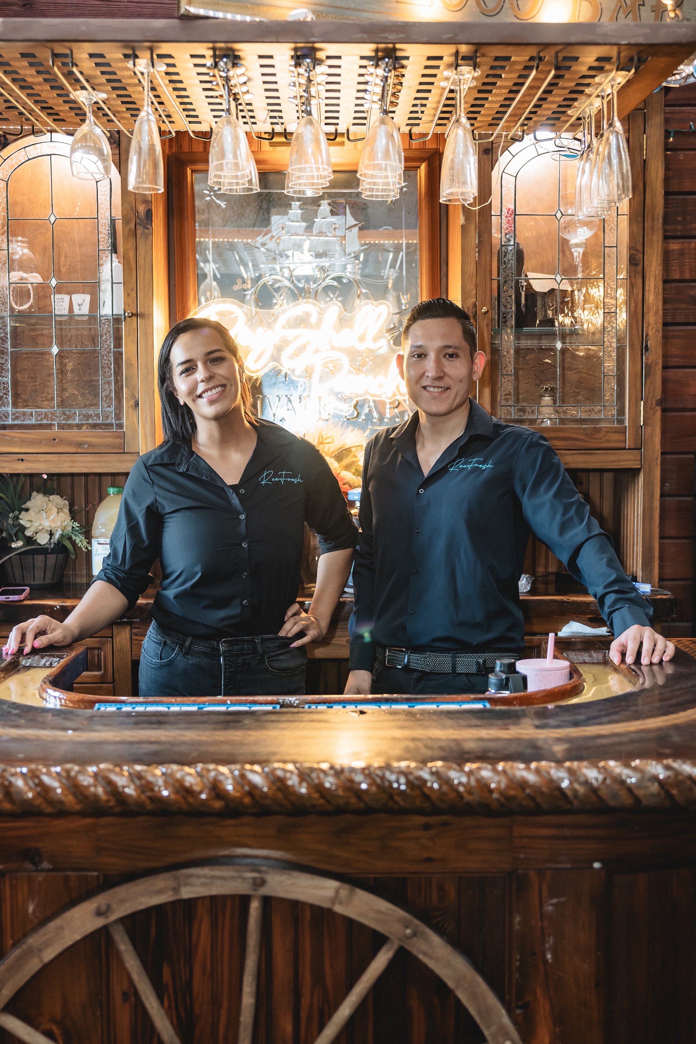 Two people smiling at a wooden bar with hanging glasses and a neon sign in the background.
