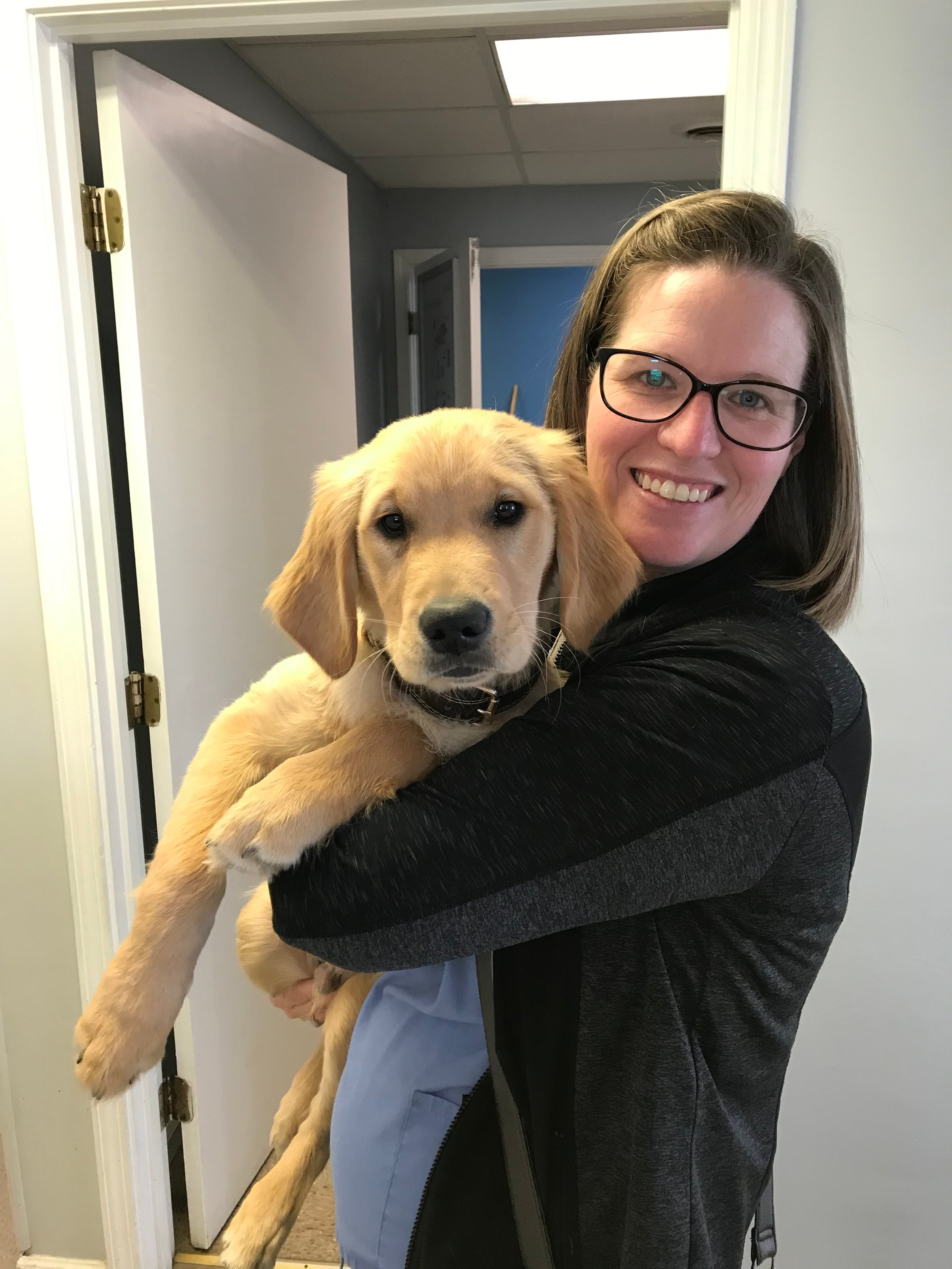 A woman wearing glasses holds a golden retriever puppy in her arms, smiling in an indoor setting.