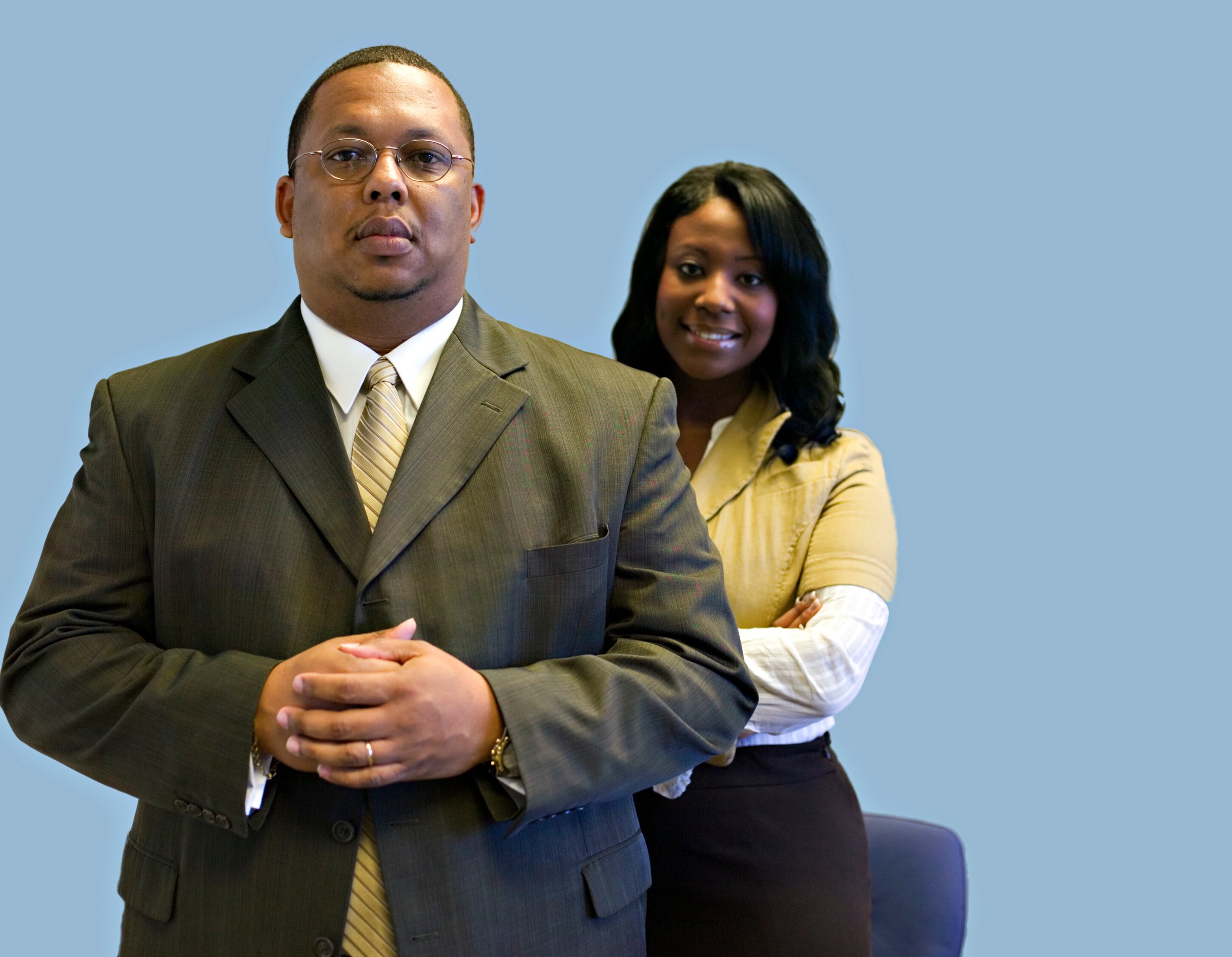 Two professionals in business attire standing against a blue background, one man in front and a woman slightly behind him.