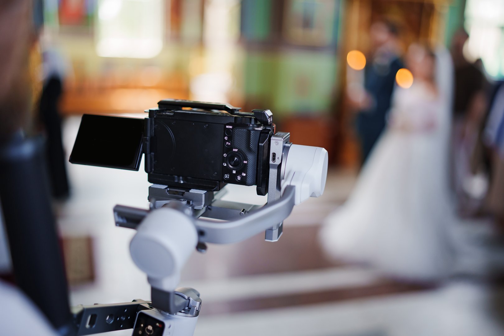 A couple stands together, exchanging vows in a stunning interior space while a videographer captures the moment with a stabilizing rig. Guests witness this joyful occasion.