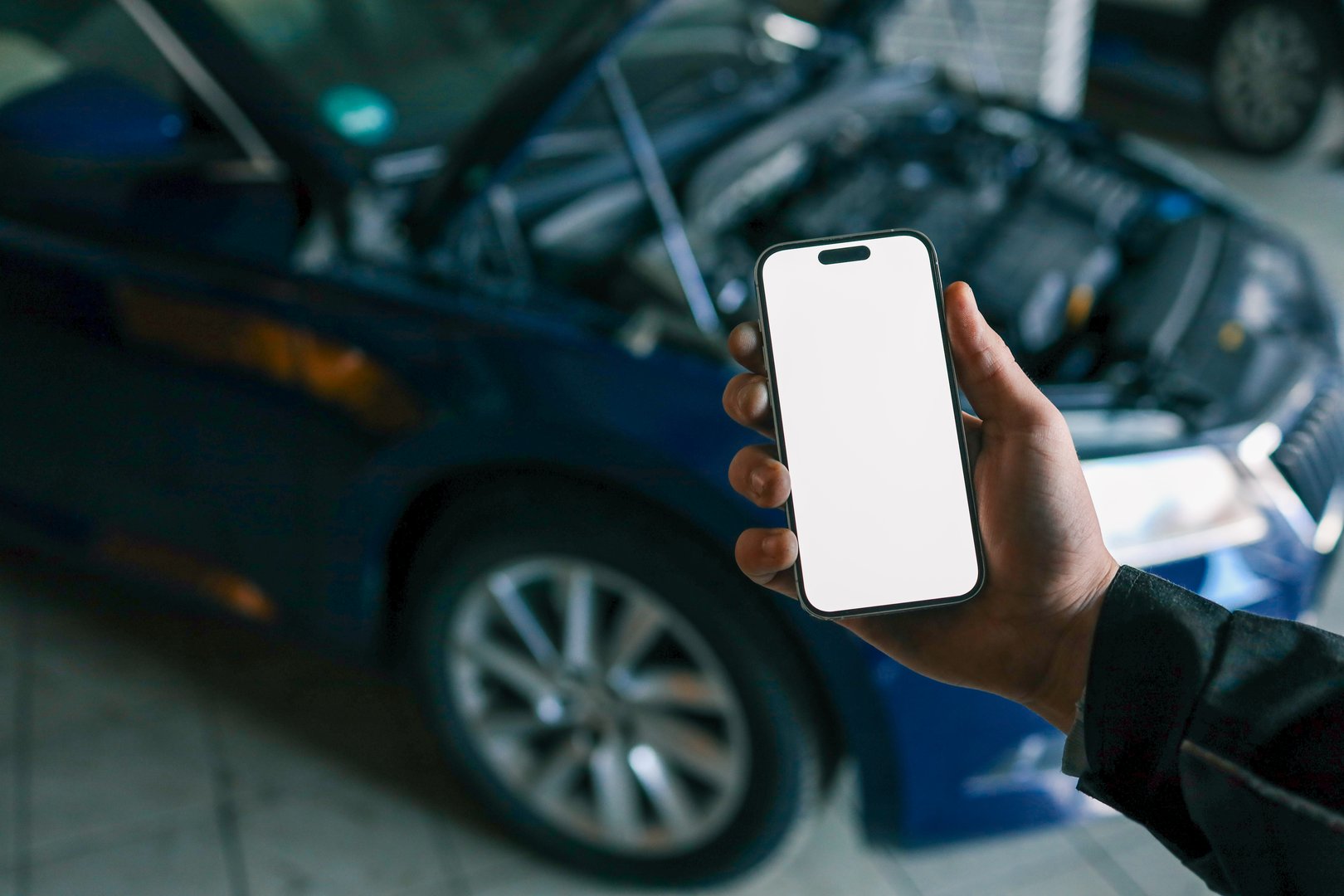 Hand holding a smartphone with a blank screen in front of an open car hood, showing the engine compartment. Concept for car diagnostics, automotive apps, or remote vehicle monitoring.