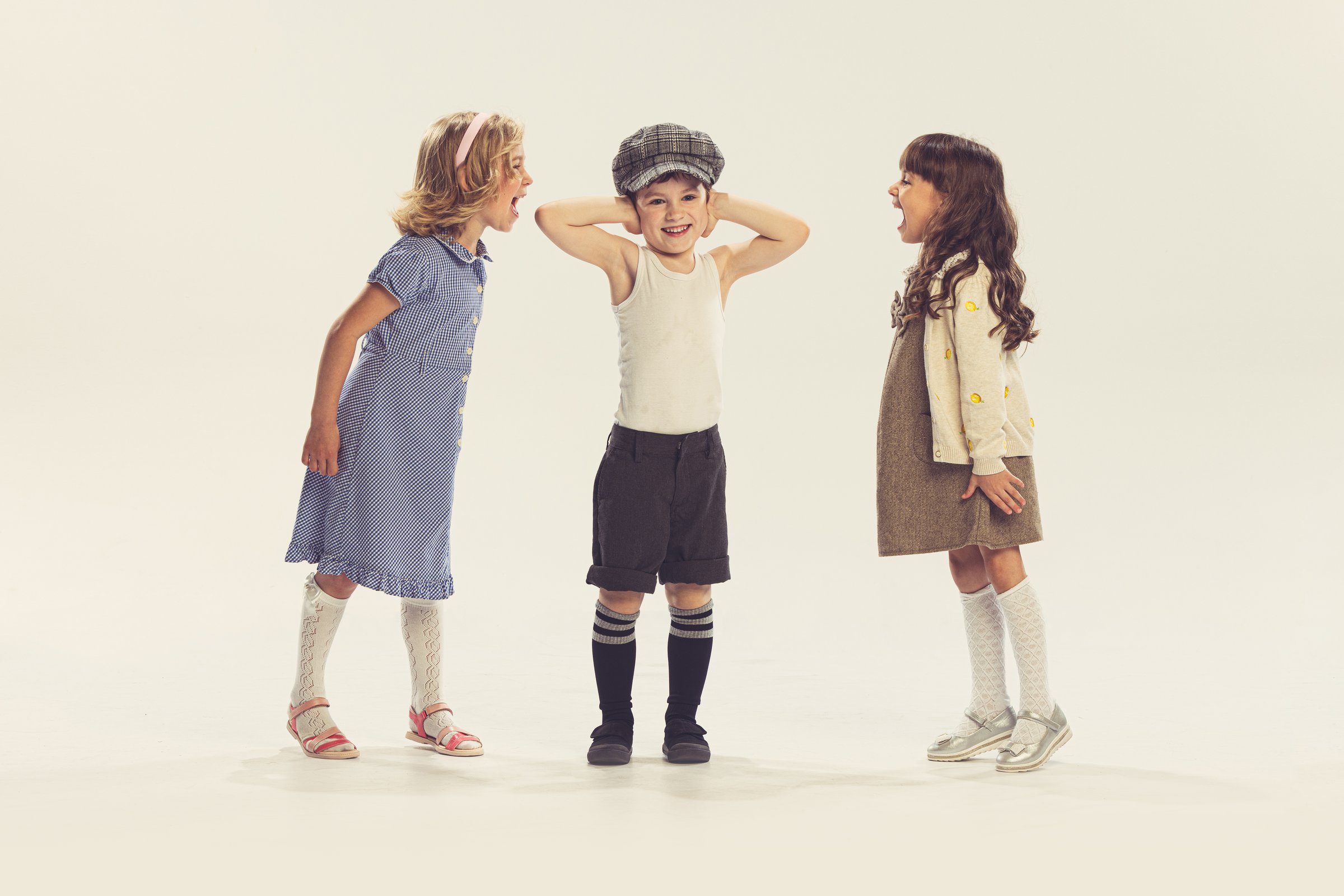 Portrait of three children playing together, girls screaming at boy covering ears isolated over grey studio background. Concept of childhood, friendship, family, fun, lifestyle, retro fashion