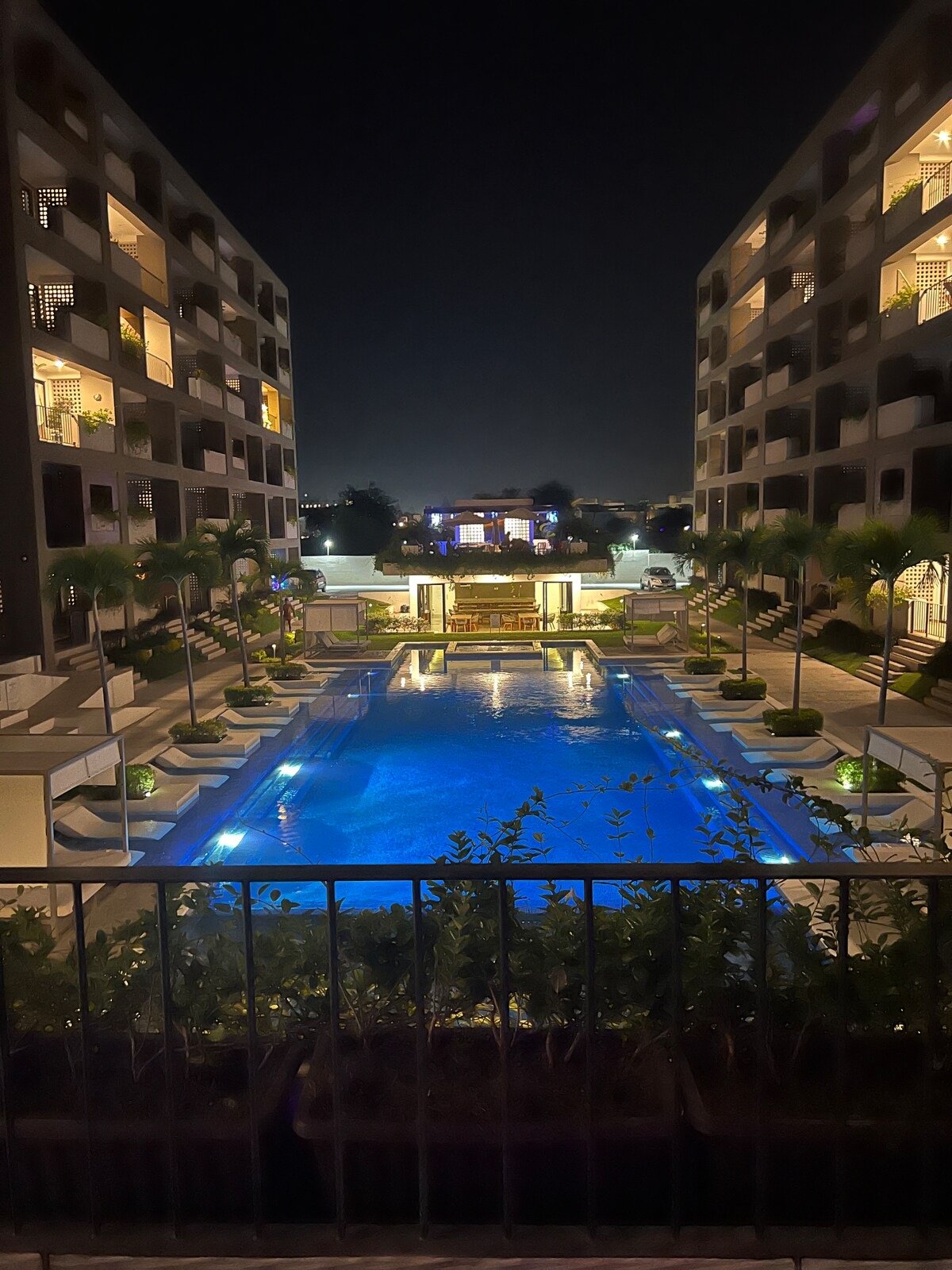 Night view of an illuminated swimming pool between two modern apartment buildings, surrounded by palm trees.