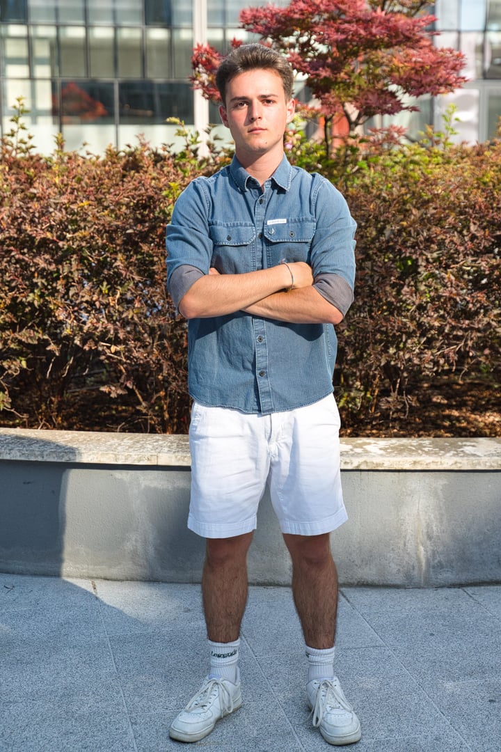 Young person standing on a wooden porch, wearing a black shirt and beige vest, with greenery and buildings in the background.