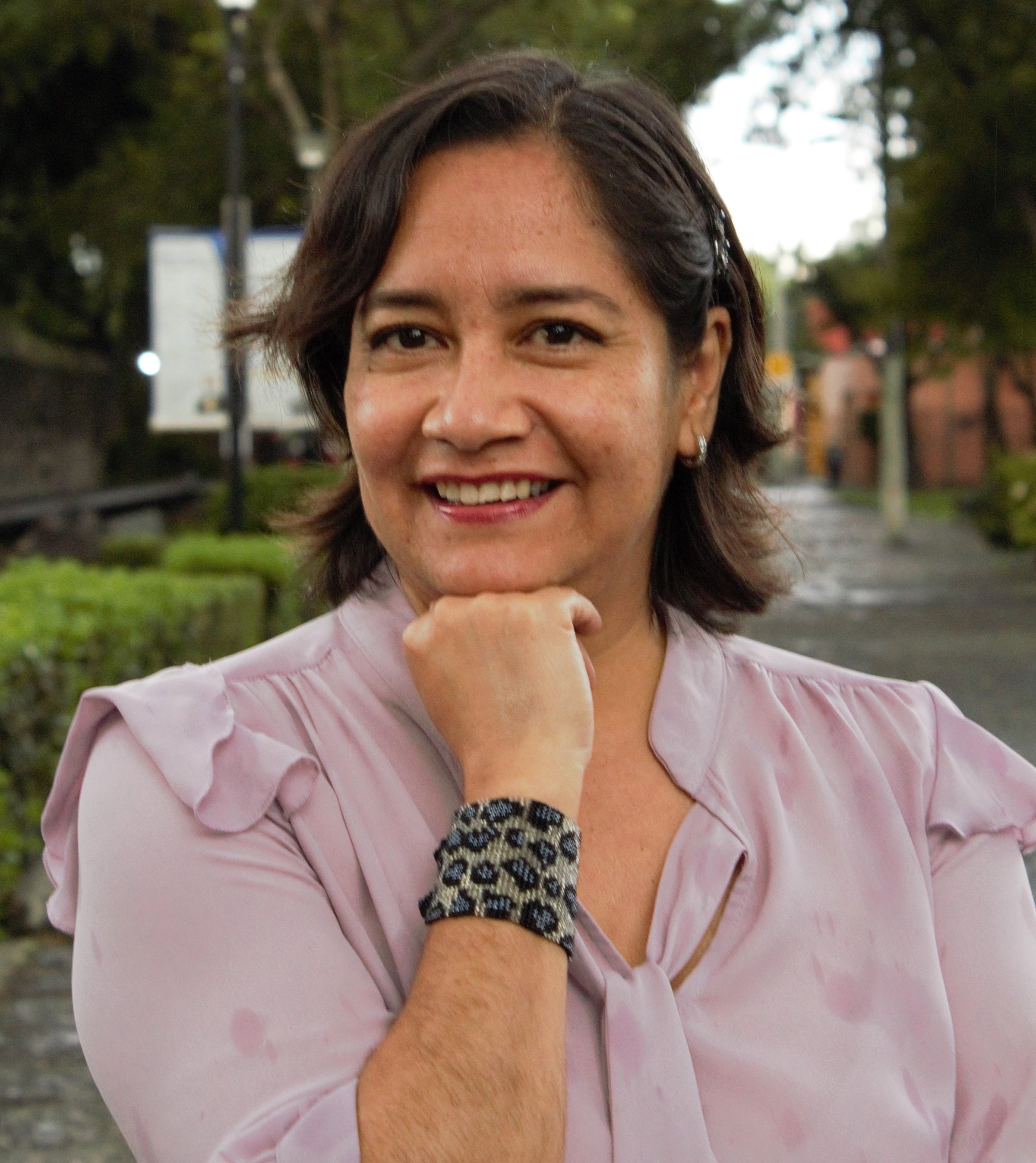 Smiling woman in a light purple blouse, with short dark hair and a beaded bracelet, posing outdoors in a park setting.