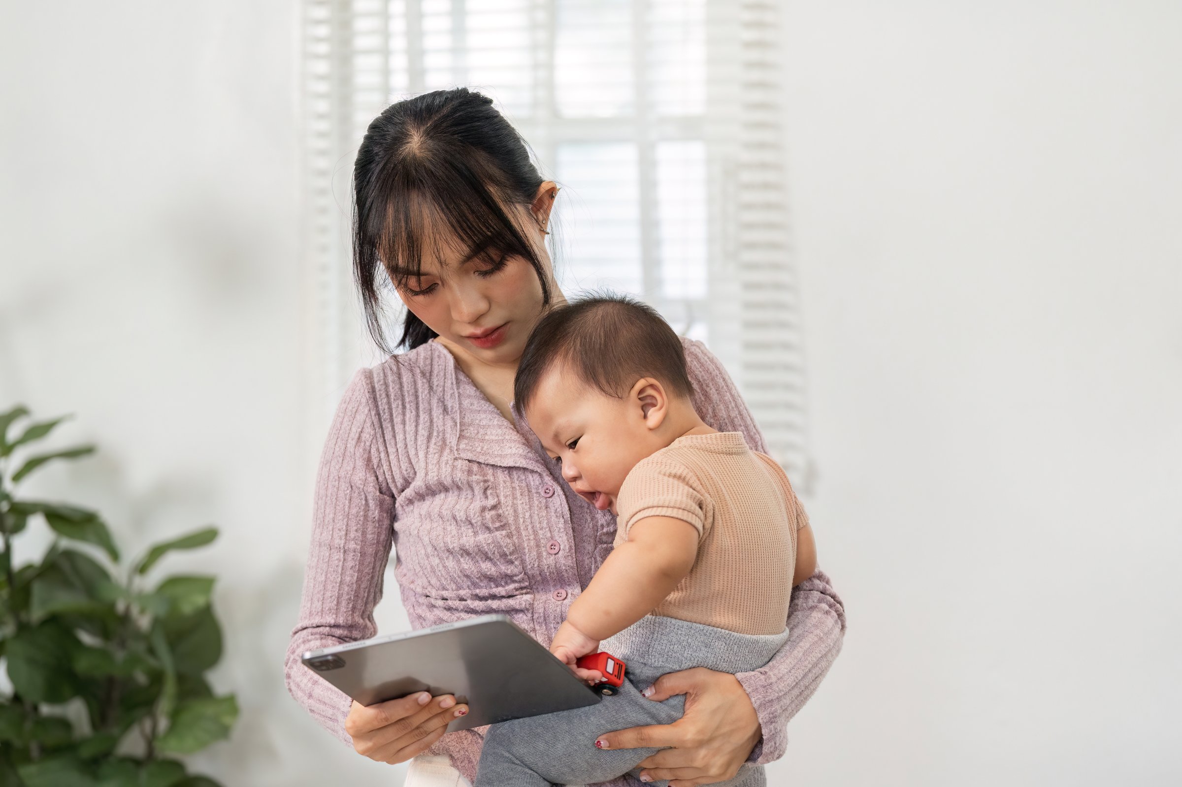 A caring mother holding her baby boy close while using a tablet in a cozy living room filled with natural light.