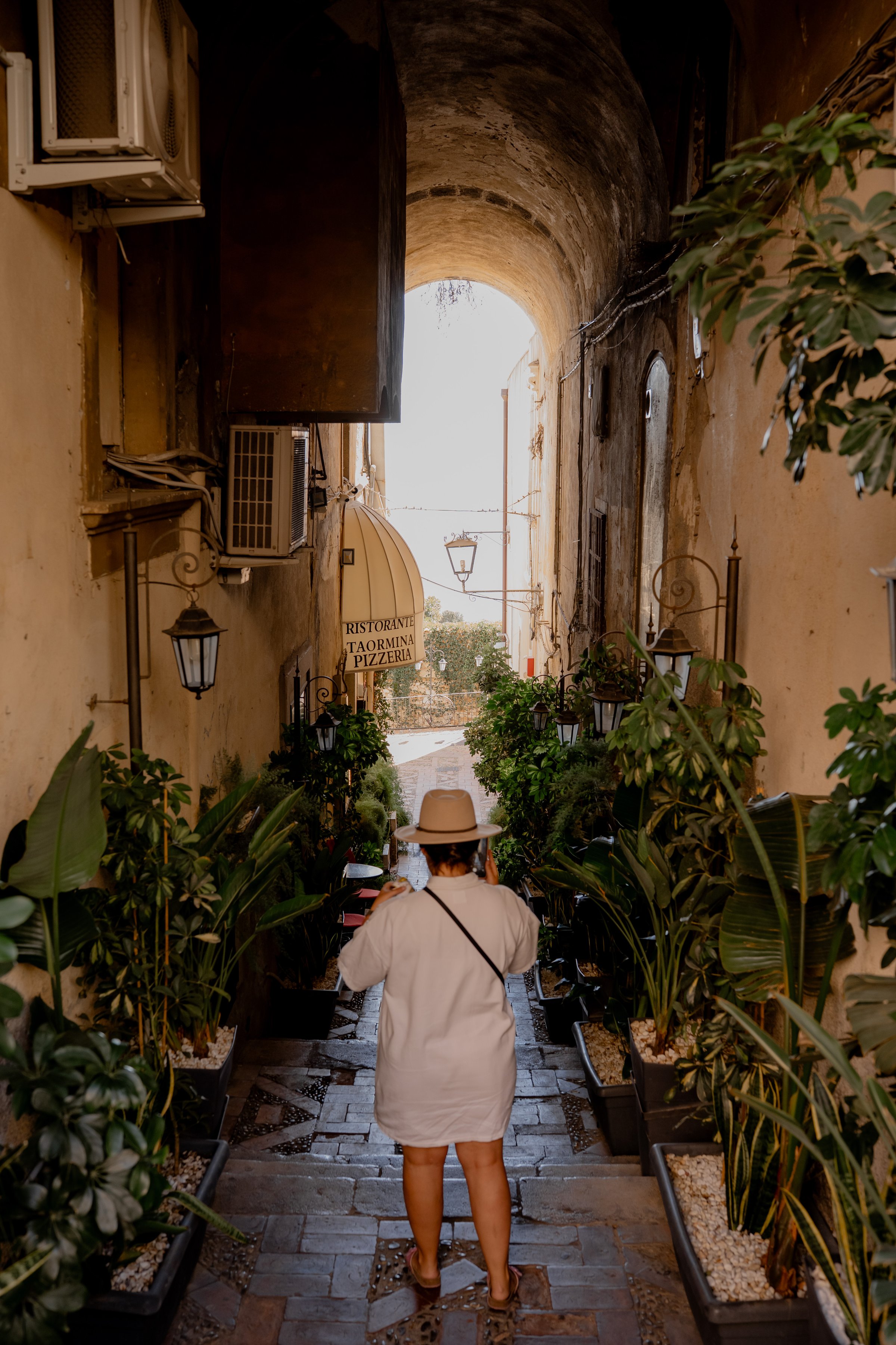 A traveler descends a narrow stone staircase under a historic archway in Taormina, Sicily. Lush plants, warm aged walls, and a view toward the street and restaurant sign create an atmospheric Mediterranean editorial travel scene.