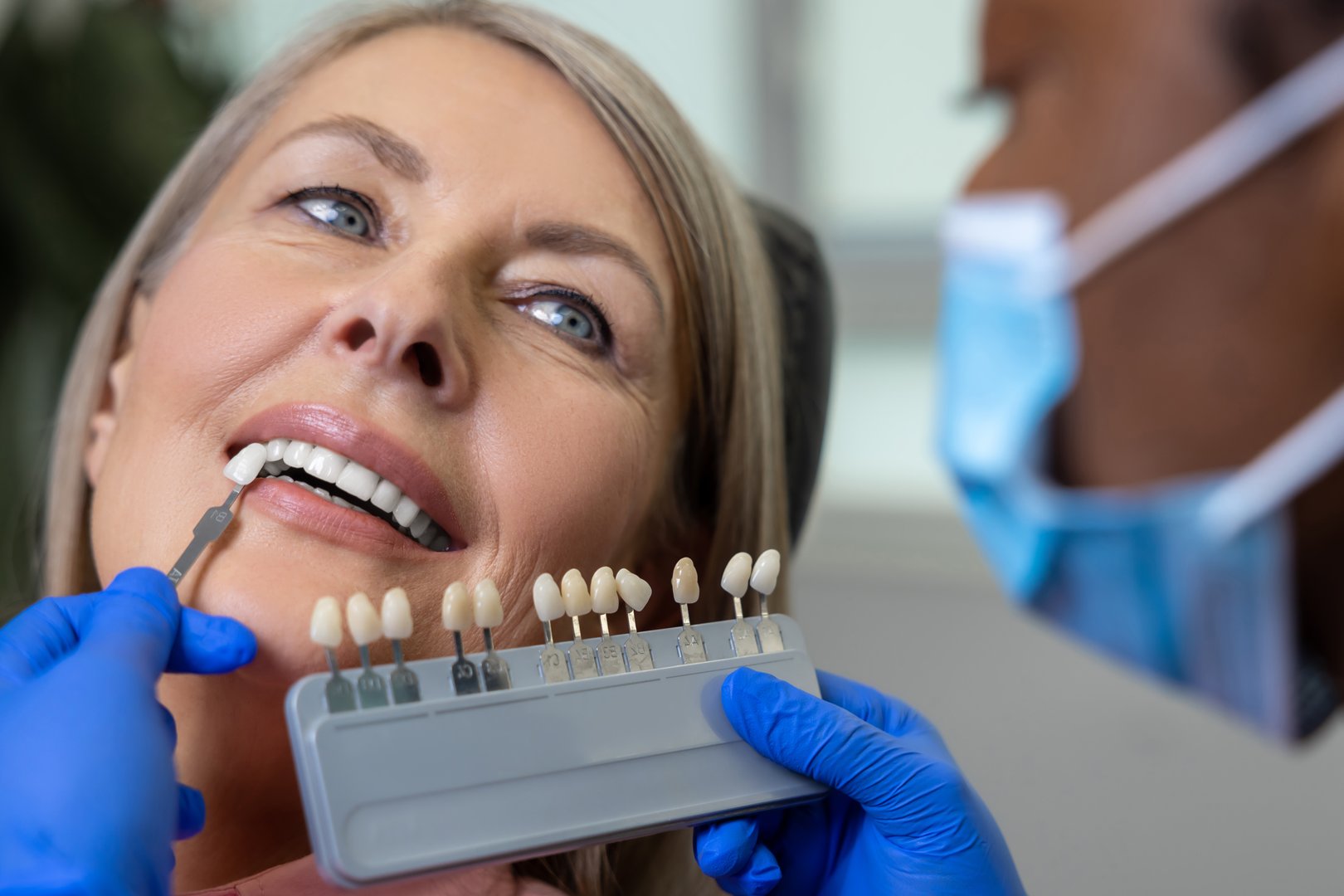 Smiling woman patient in dentist's seat while doctor orthodontist choosing the shade of dental veneer according to color chart