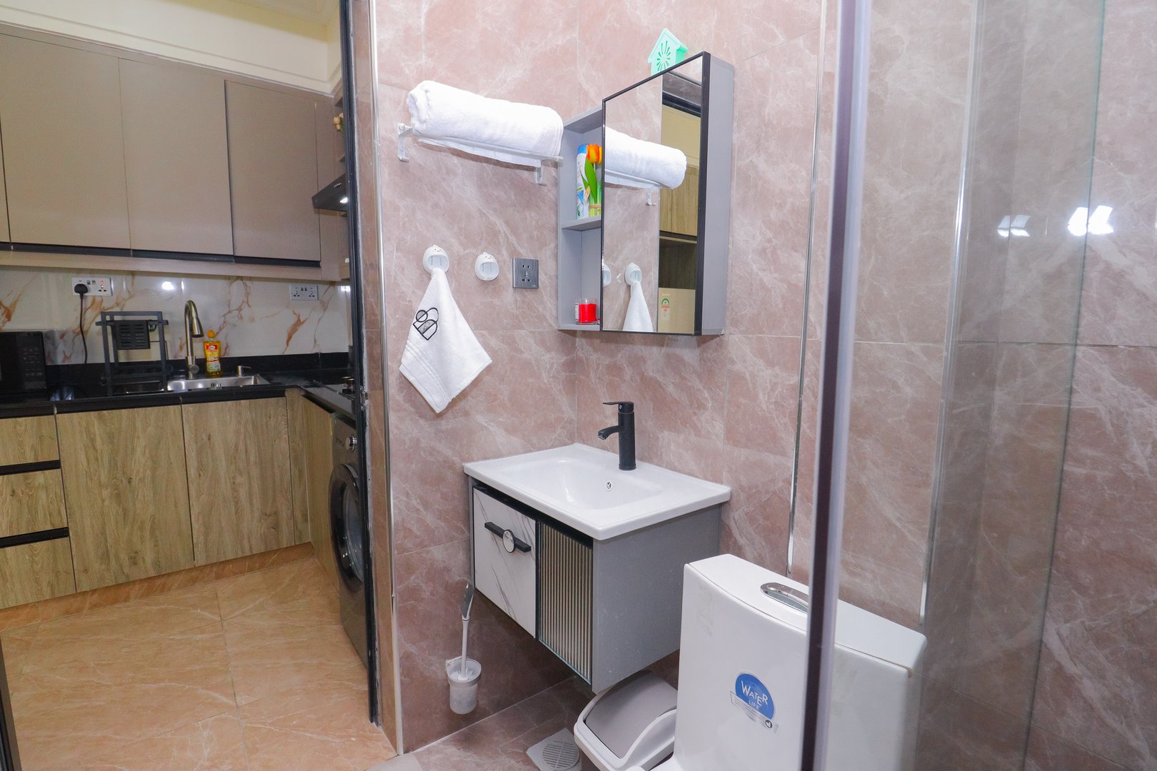 Modern bathroom with a glass shower, sink, and toilet next to a compact kitchen with light wood cabinets.