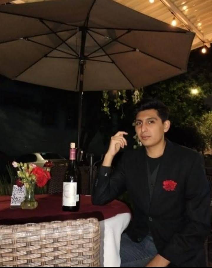 Man sitting at an outdoor table with a bottle of wine and flower arrangement under an umbrella, wearing a black jacket.
