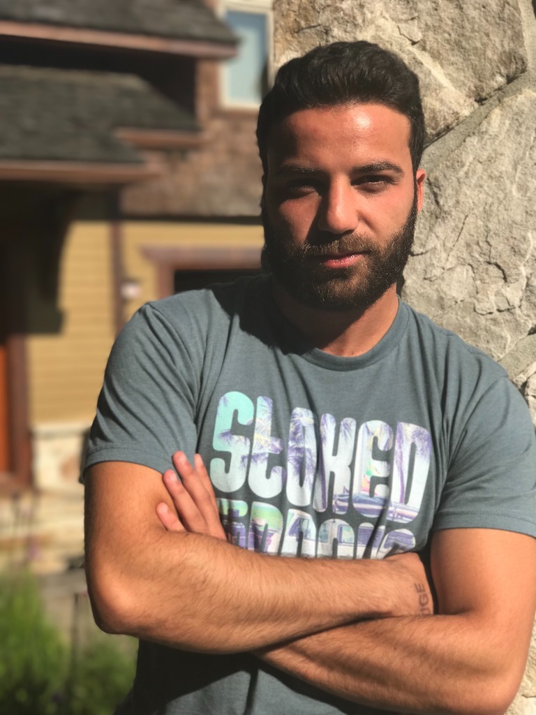 A man with a beard and mustache stands outside with arms crossed, wearing a blue T-shirt on a sunny day.