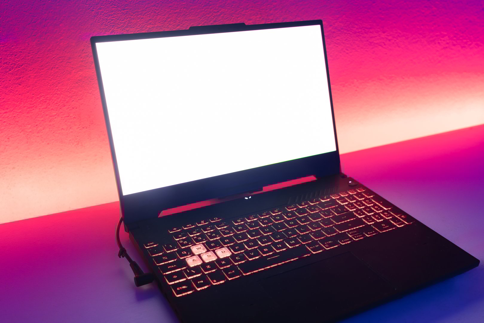 Close-up of a gaming laptop with a blank white screen and illuminated keyboard in a vibrant LED-lit environment. The pink and purple gradient background creates a modern and tech-inspired atmosphere