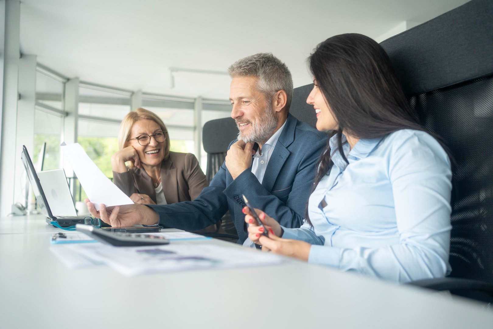 Three happy middle-aged professionals collaborating on laptop for financial project in office.