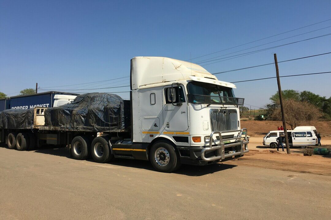 A white semi-truck with a trailer loaded with covered cargo parked on the side of a road under a clear blue sky.