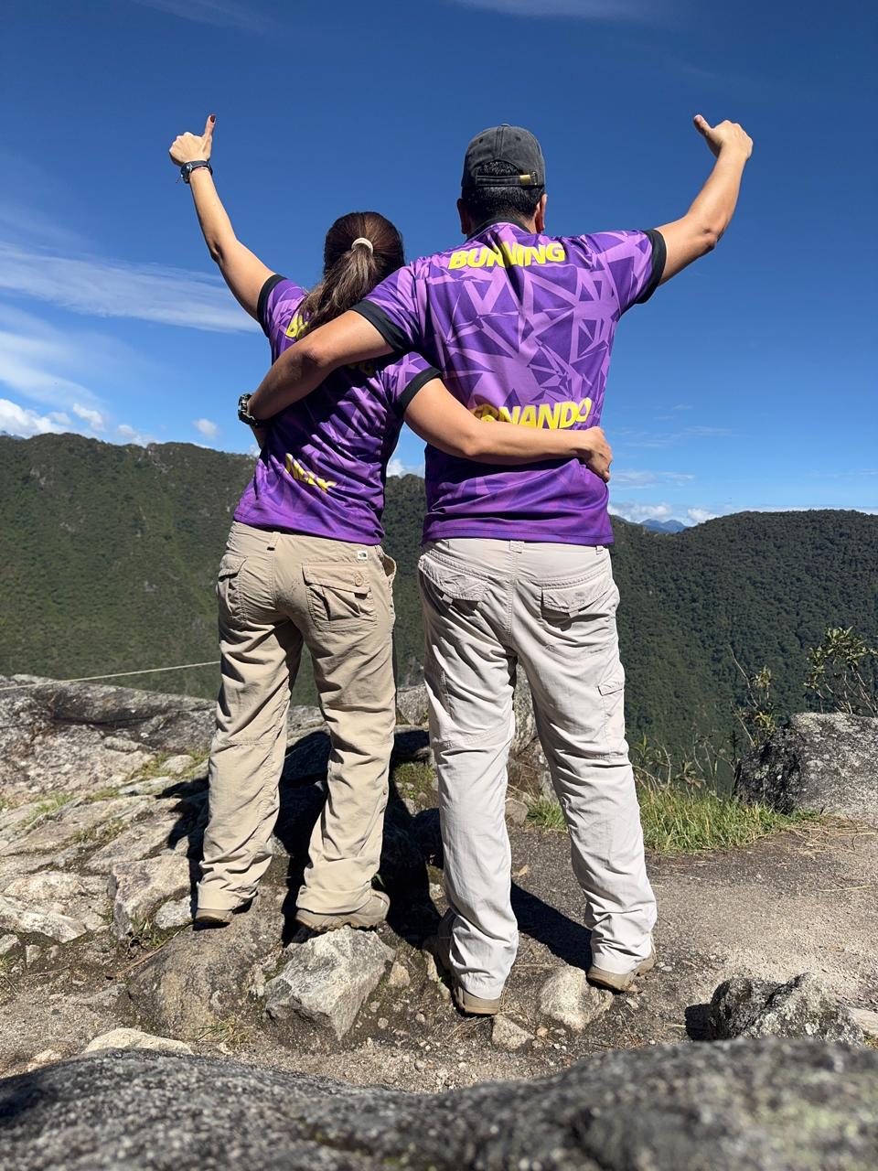 Two people in matching purple shirts overlook a scenic mountain view, raising their arms in celebration.