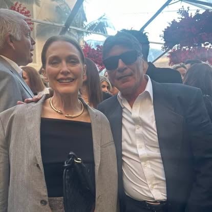 A man wearing sunglasses and a woman smiling, both dressed in formal attire, stand together at an indoor event.