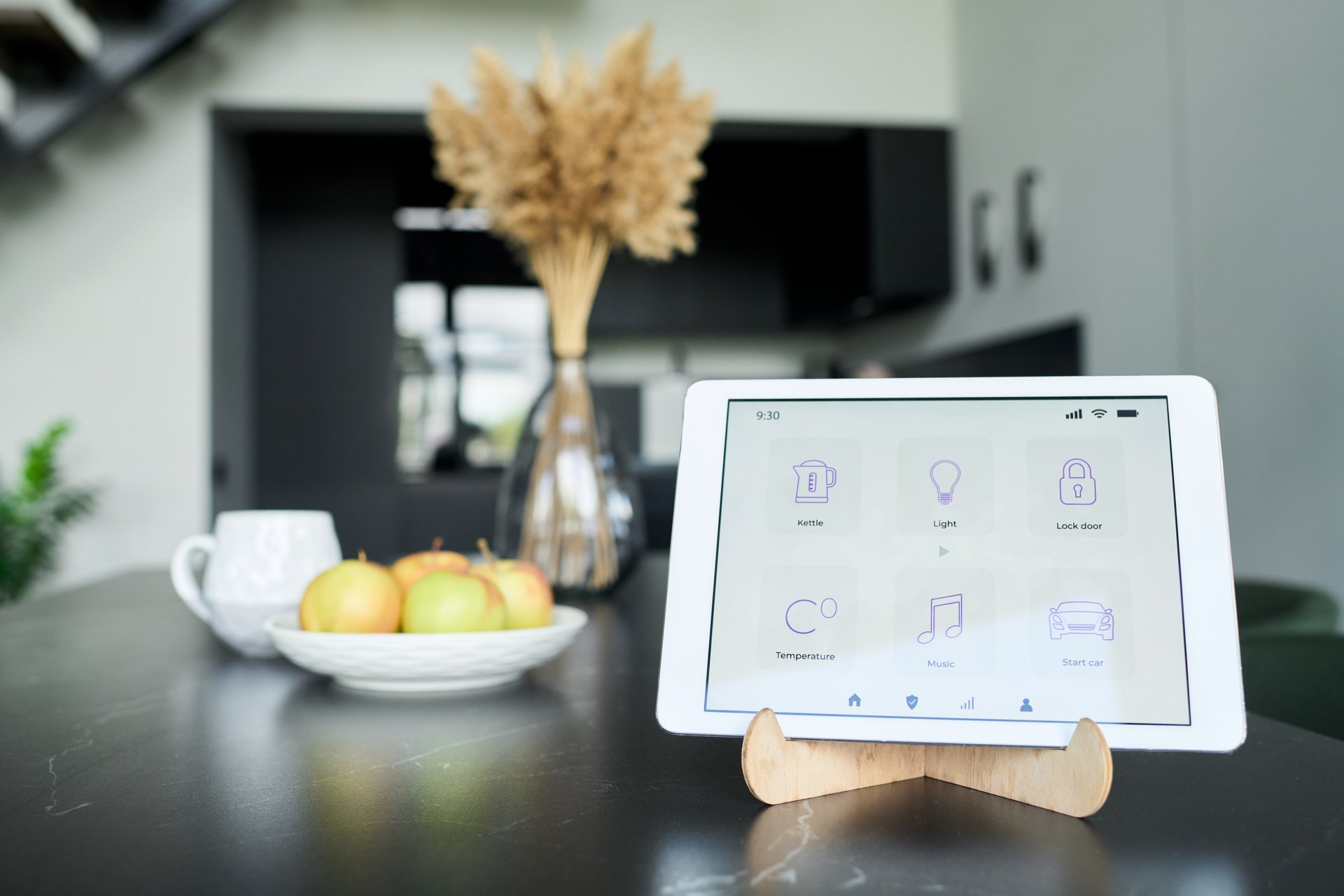 Home automation control tablet situated on kitchen counter surrounded by fresh apples in bowl and dried flowers in vase. Light-filled kitchen creating serene setting