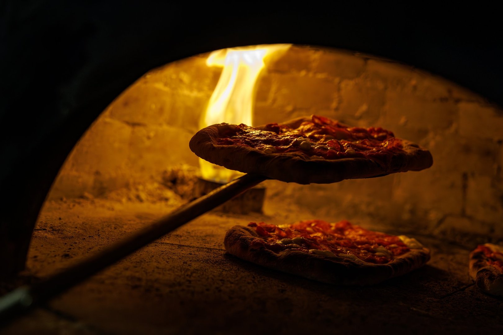 Pizza being lifted with a metal peel from a hot wood-fired oven with bright flames inside.