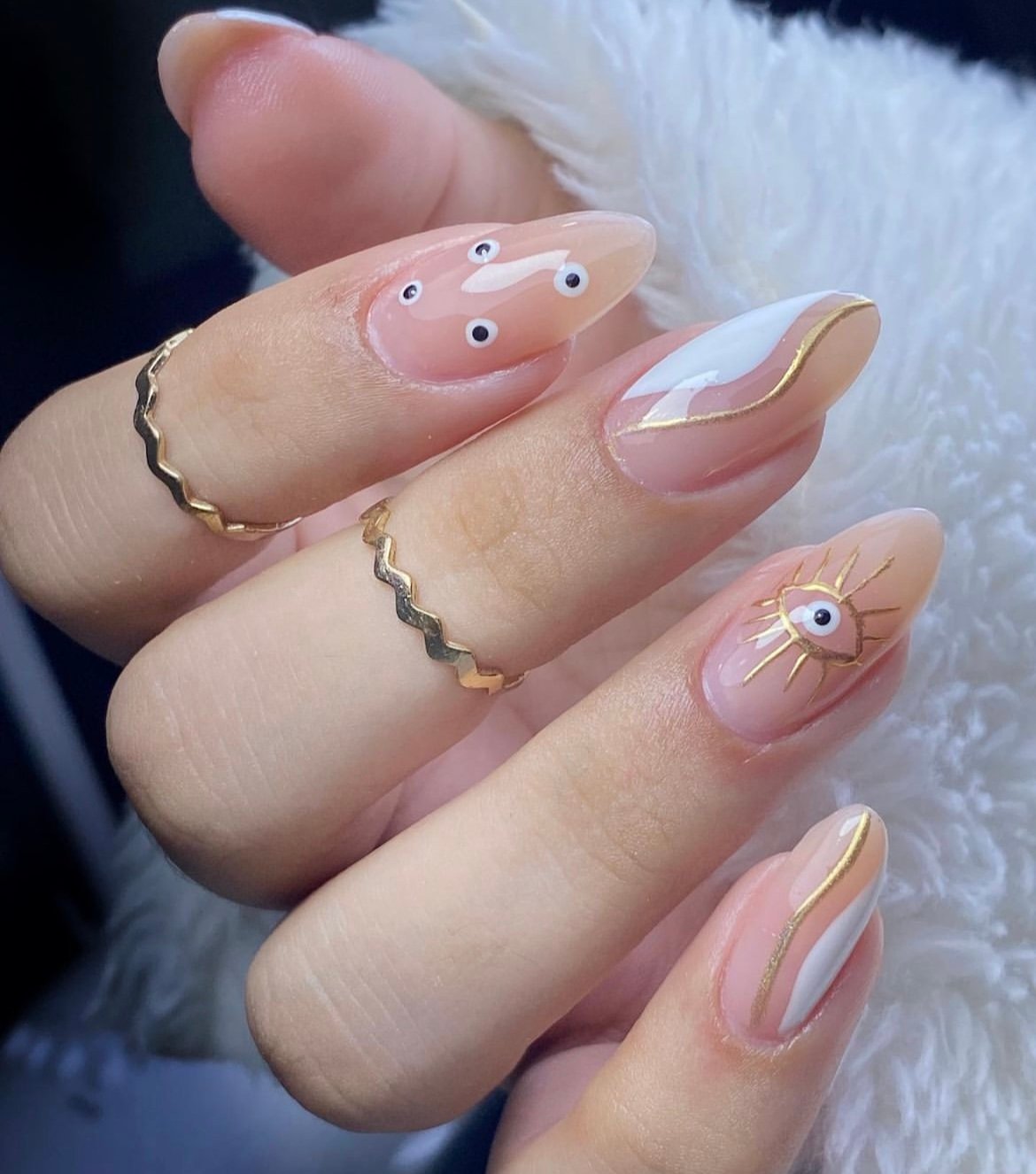 Hand with pink and white nail art featuring eyes and golden accents, wearing gold rings on fingers.