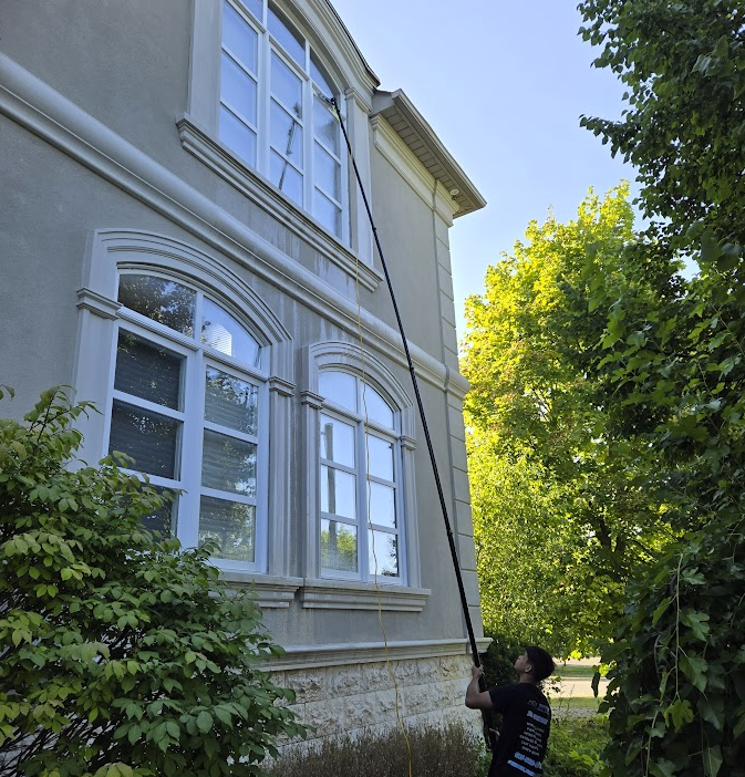 Person cleaning high windows of a house using an extendable pole, surrounded by greenery and trees.
