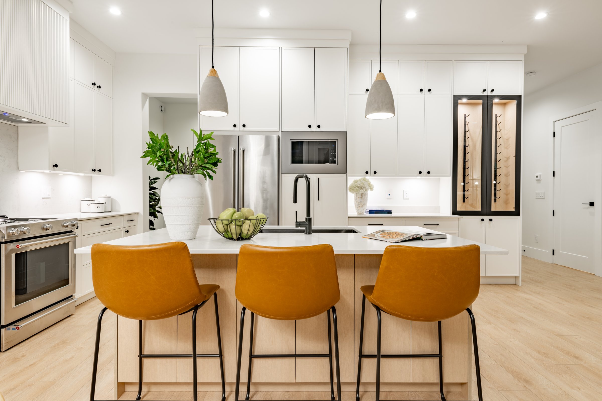 Kitchen and dining room in a newly constructed residential home, photography was done at twilight showing blue hour skies through the windows. Kitchen island has 3 carmel coloured bar stools and a white granite countertop. Cabinets are a light coloured wood with a built in wine fridge, there are stainless steel appliances. The floor is a light hardwood and the dining table is an oval shape with curved dining chairs.