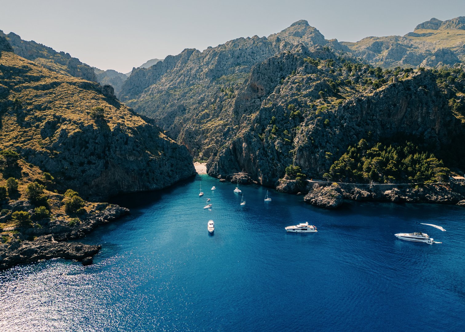 Aerial view of yachts and boats anchored in a secluded bay surrounded by towering rocky cliffs and rugged mountain landscape, with clear blue waters under the bright sky