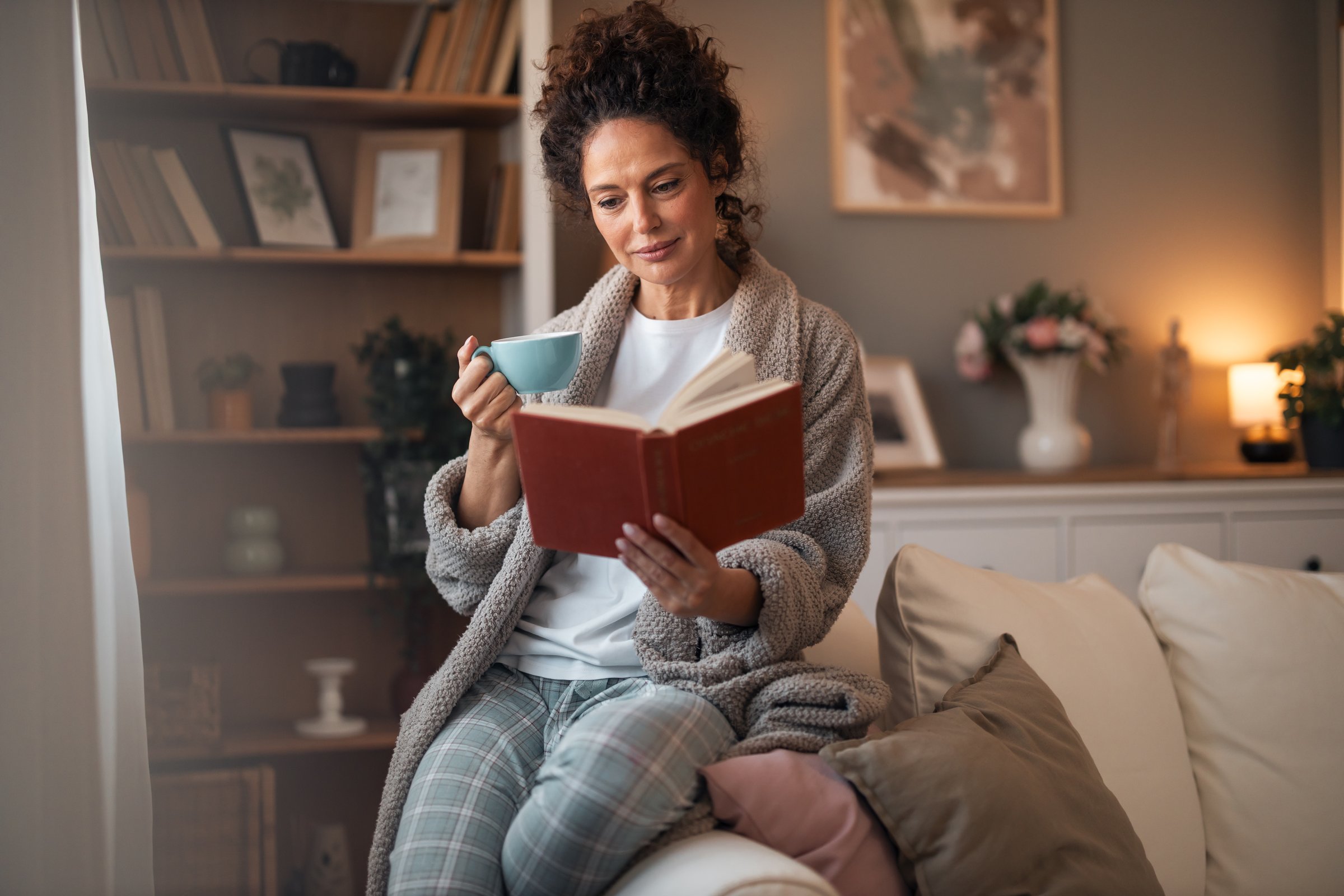 A woman sits on a sofa at home, reading a red hardcover book and sipping from a blue cup. Soft lighting, cozy robe, and calm, intimate atmosphere, perfect for home comfort.