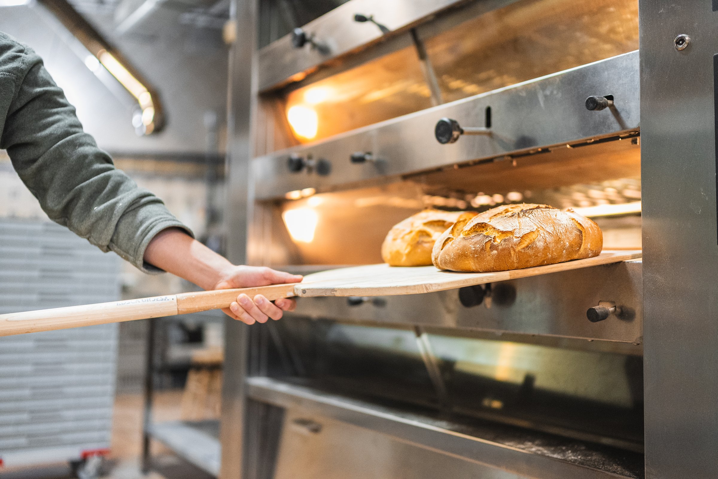 Baker removing two loaves of sourdough bread from a professional oven using a wooden peel in a bakery
