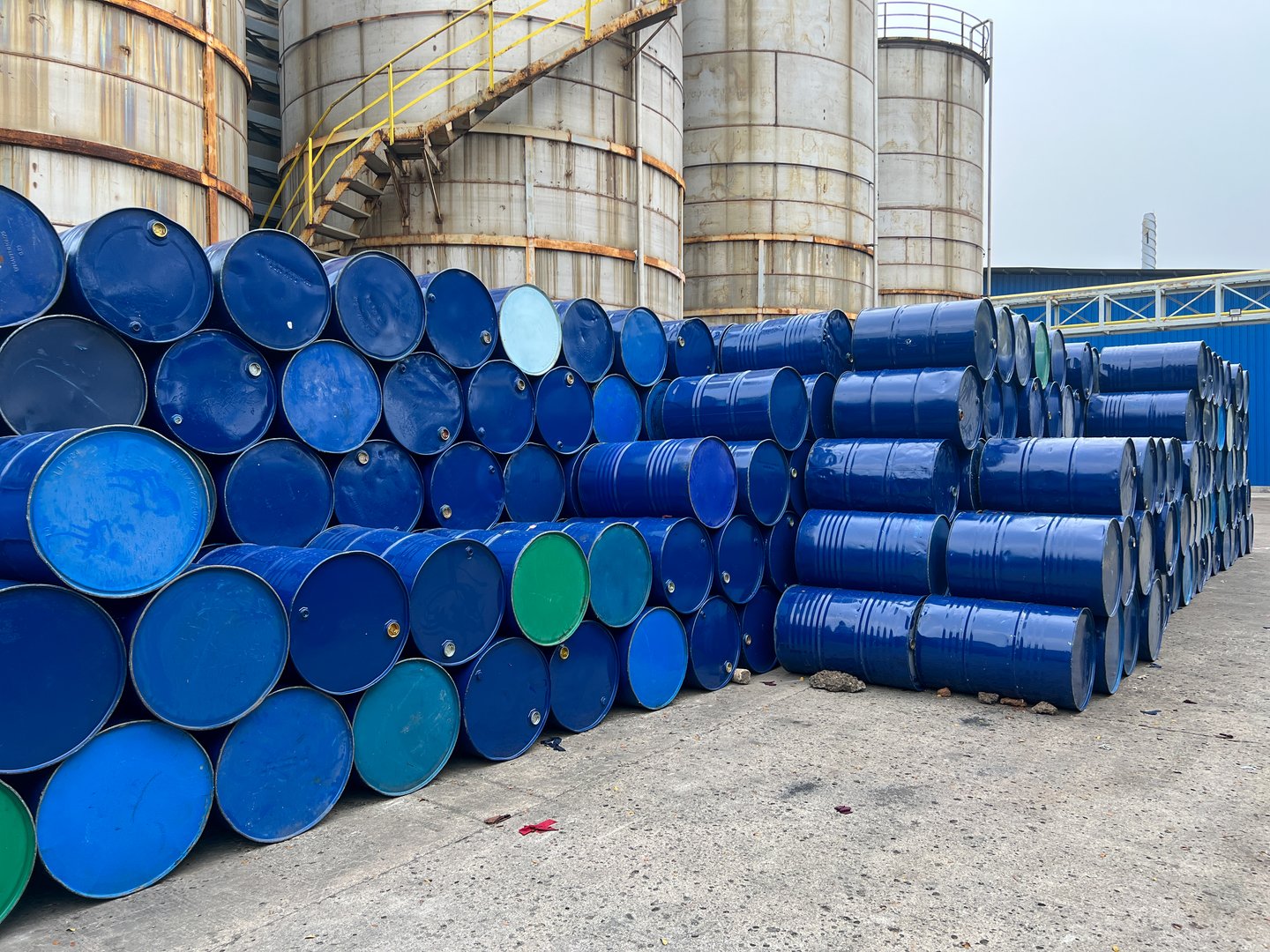 A pile of blue metal barrels stored outdoors at an industrial facility against a factory backdrop.