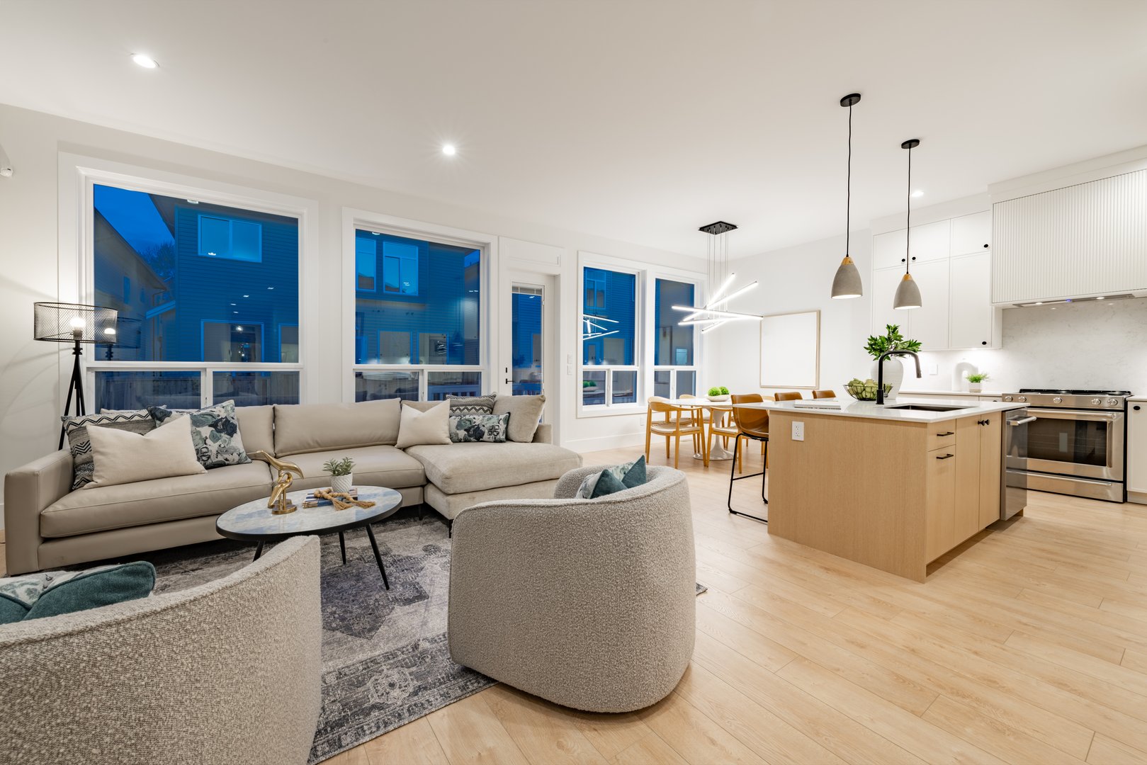Living room interior of a new construction home in a residential neighborhood, the photos show the blue hour twilight through the windows. The family home features comfortable upholstered furniture a fireplace and simple decor in an open concept space with a view from the living room to the kitchen and dining room. Hardwood floor has an area rug in the middle.
