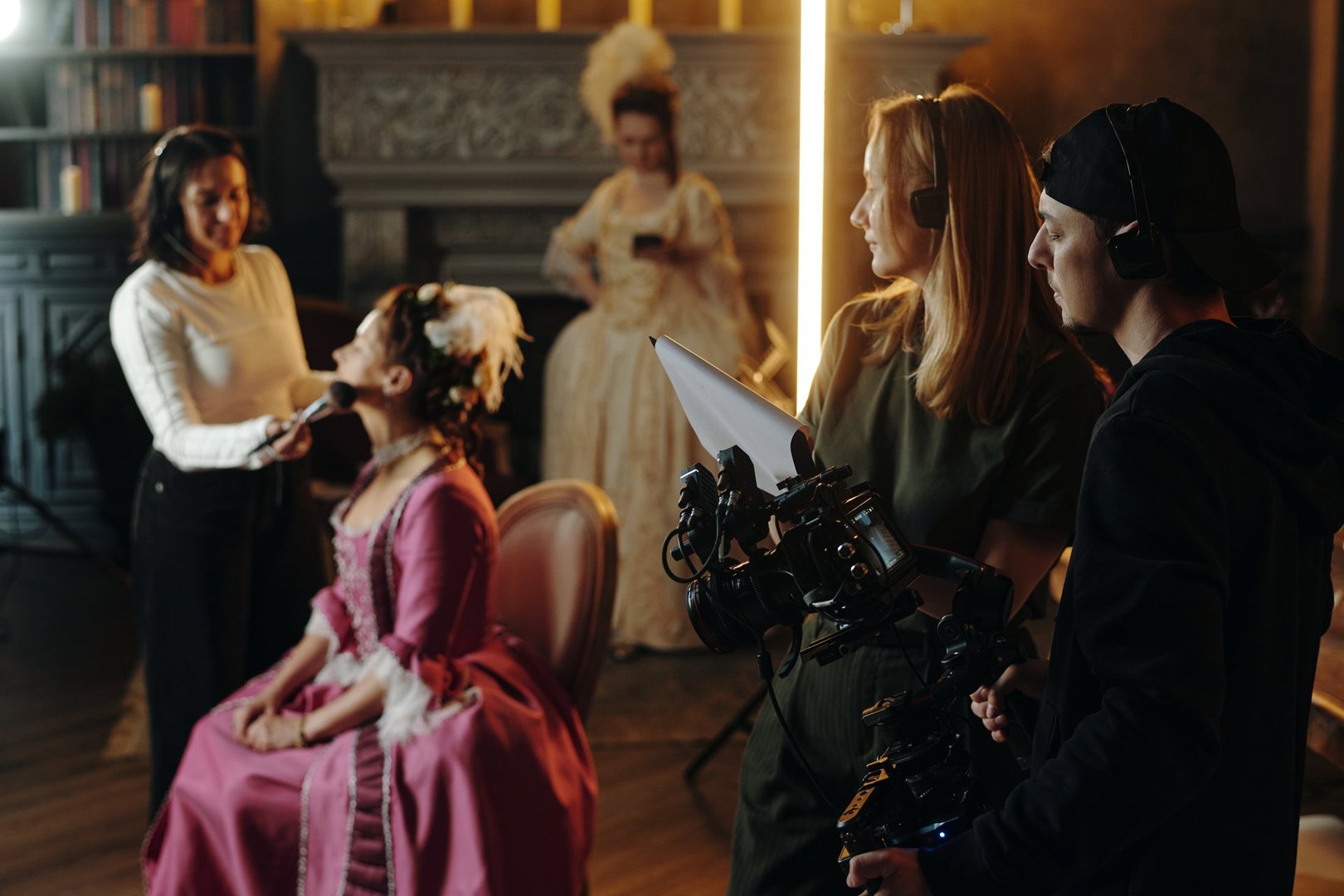 Caucasian young adult woman sitting in chair receiving makeup from another woman while film crew preparing camera on Baroque Renaissance set