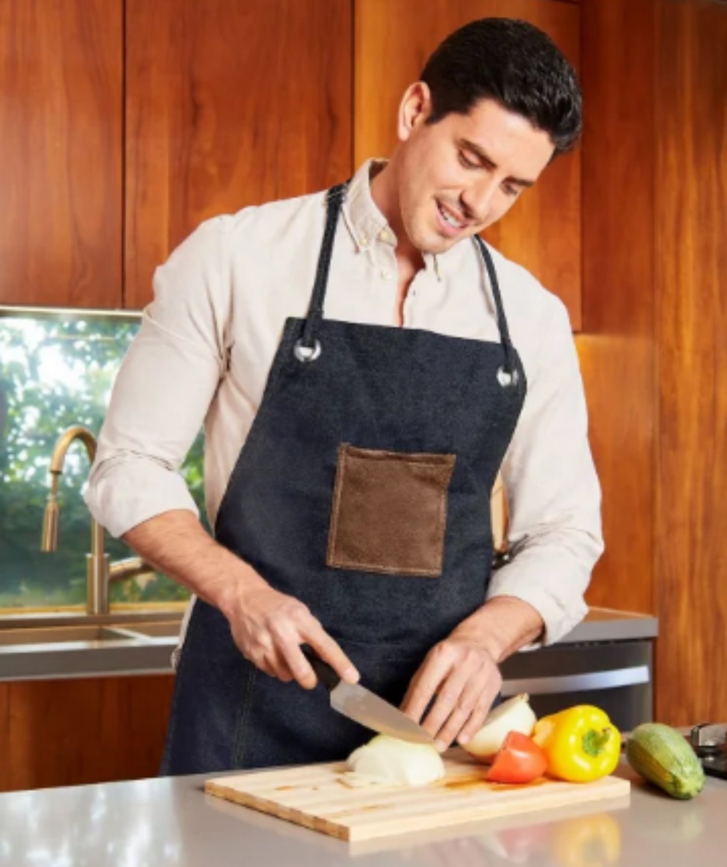 Man in a kitchen wearing an apron, smiling and chopping onions on a cutting board with bell peppers nearby.