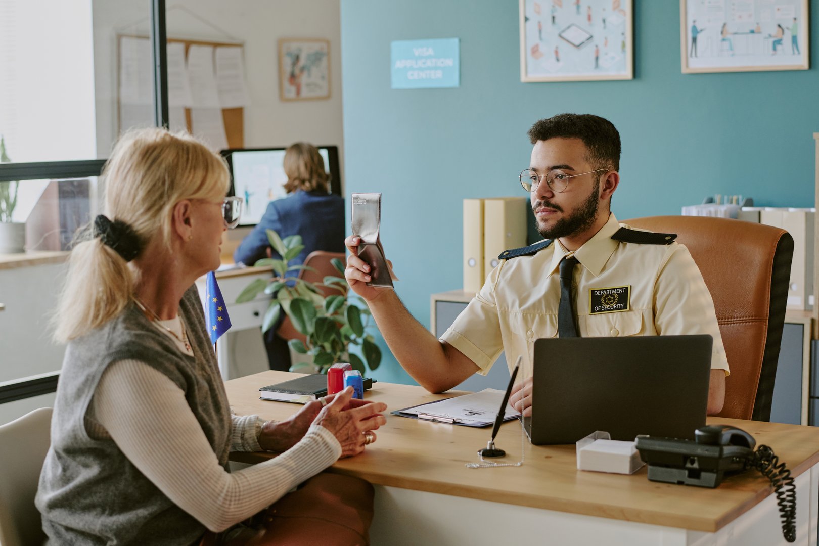 Visa officer inspecting passport at his desk in work application center while speaking with senior woman. Office setting with paperwork and computer in background