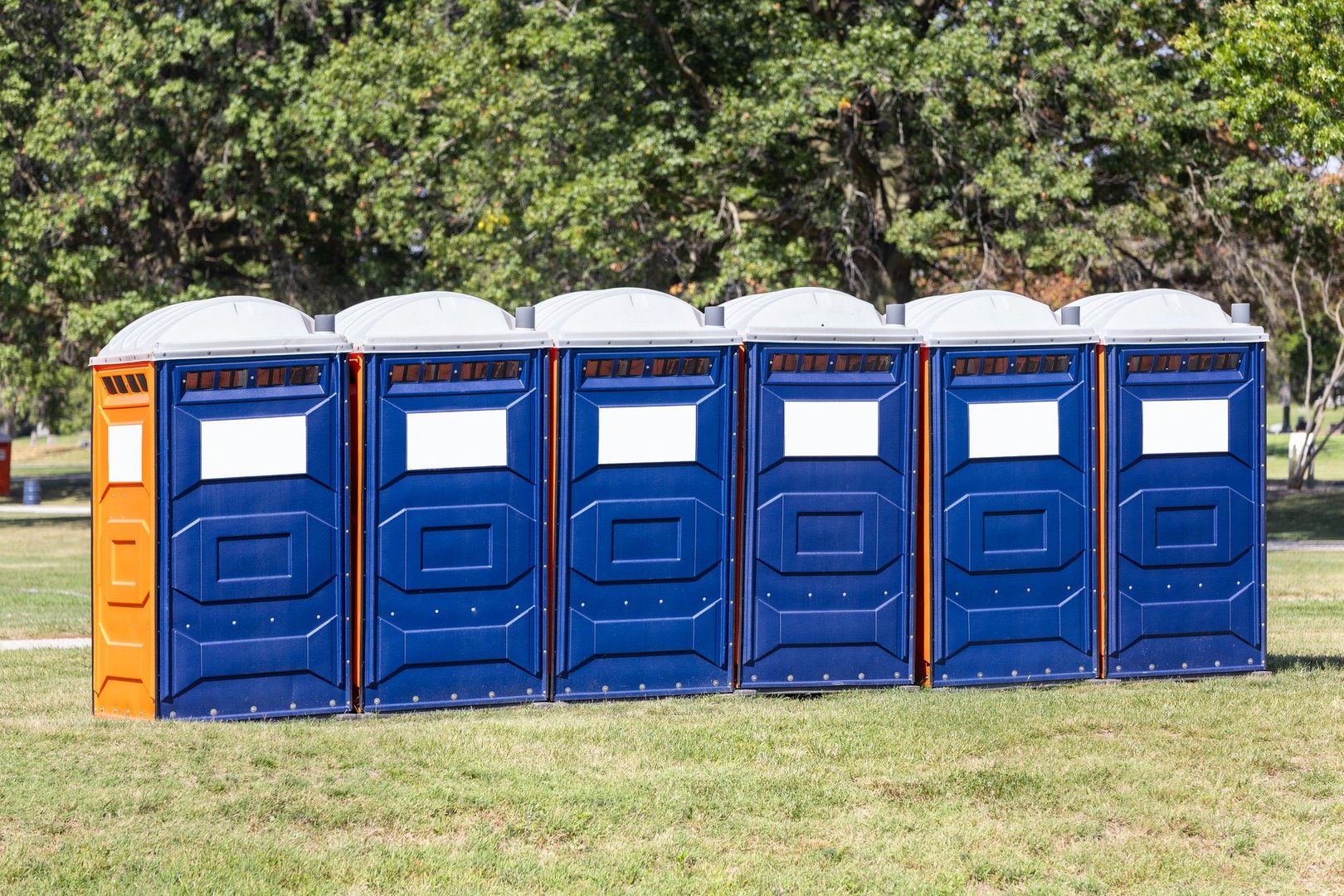 A row of portable restrooms in a park that are blue and orange with a white roof and blurred trees in the background. A blank white space for a sign or logo.