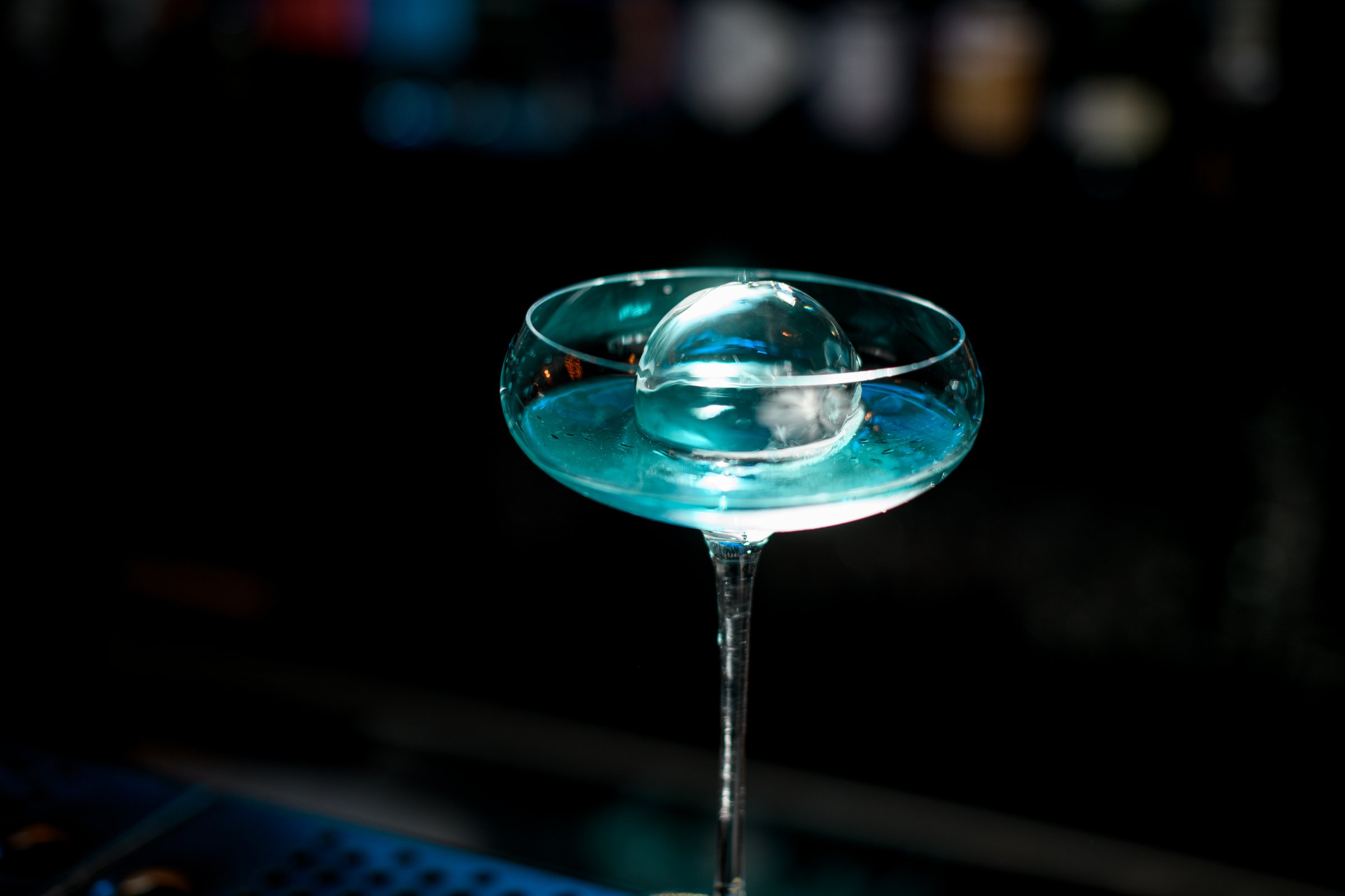 Close-up of very beautiful glass with blue drink and piece of round ice stand on bar counter.