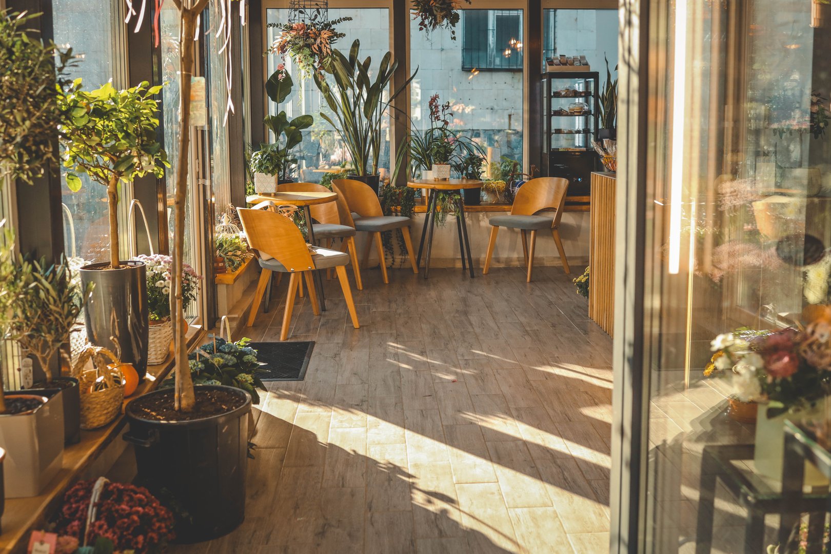 Bright and airy indoor space filled with flowers, plants, and cozy wooden furniture. Natural light streams in through large windows, creating a peaceful and inviting atmosphere.