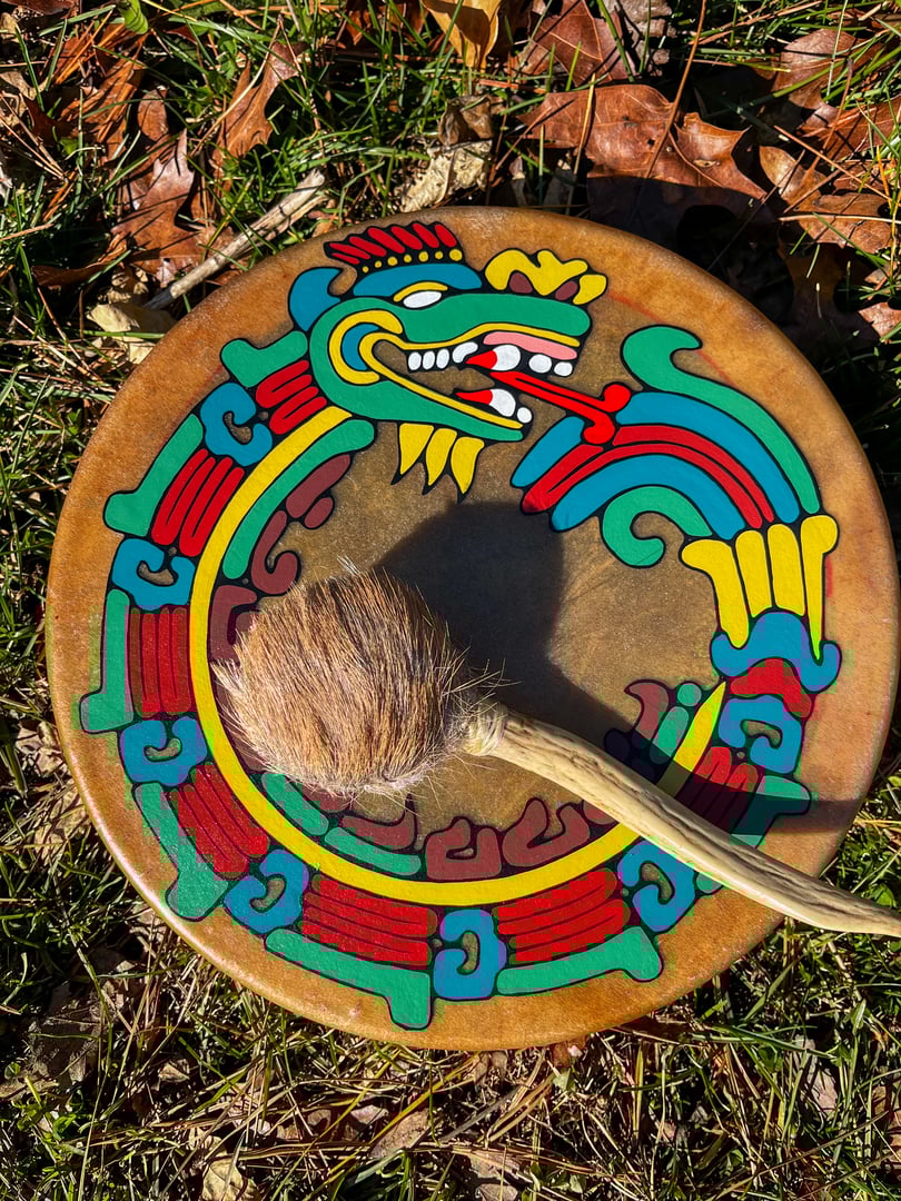 Decorative drum with colorful Aztec-inspired patterns and a drumstick on grass.