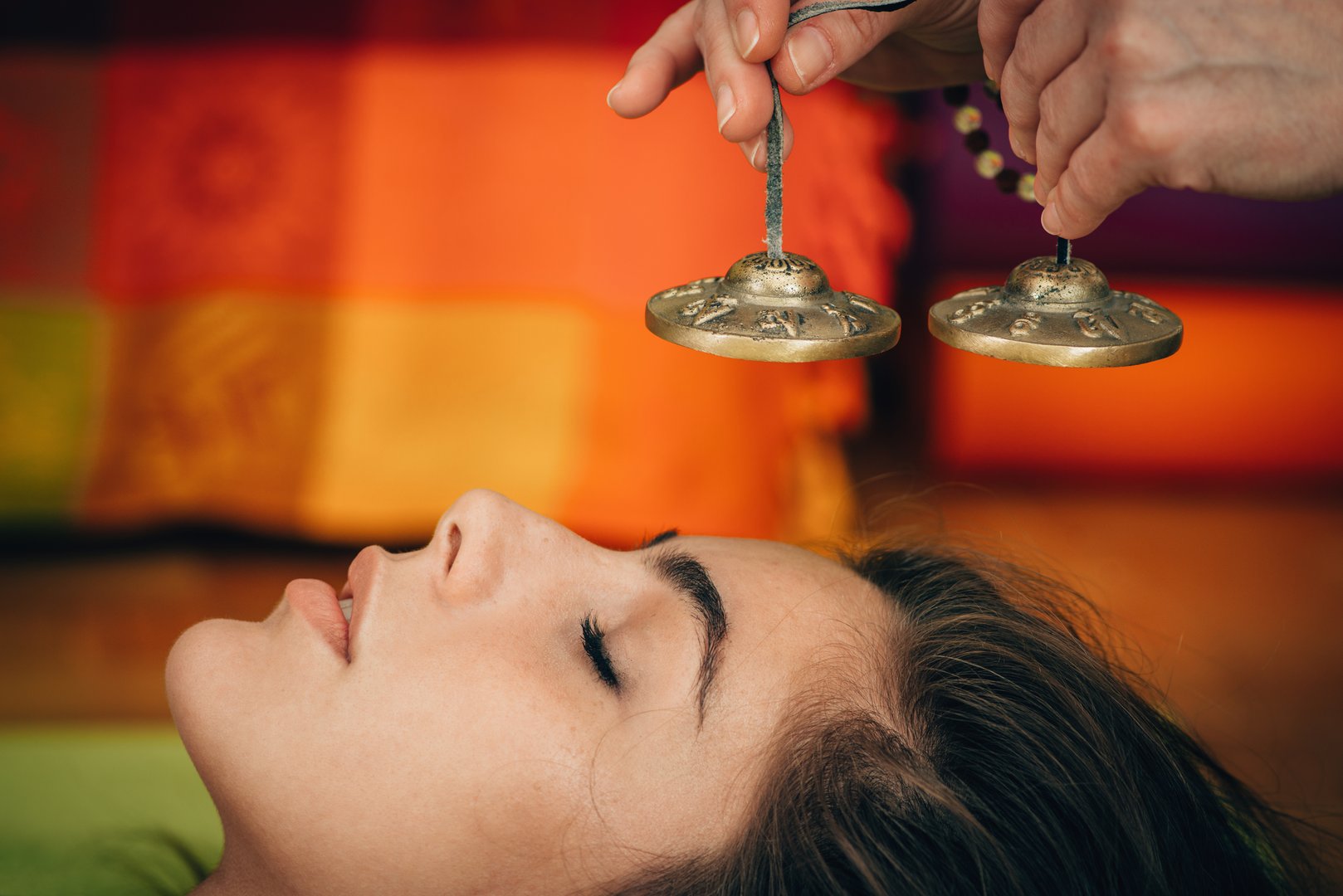 Person lying down with eyes closed as hands hold Tibetan cymbals above their face, colorful background visible.