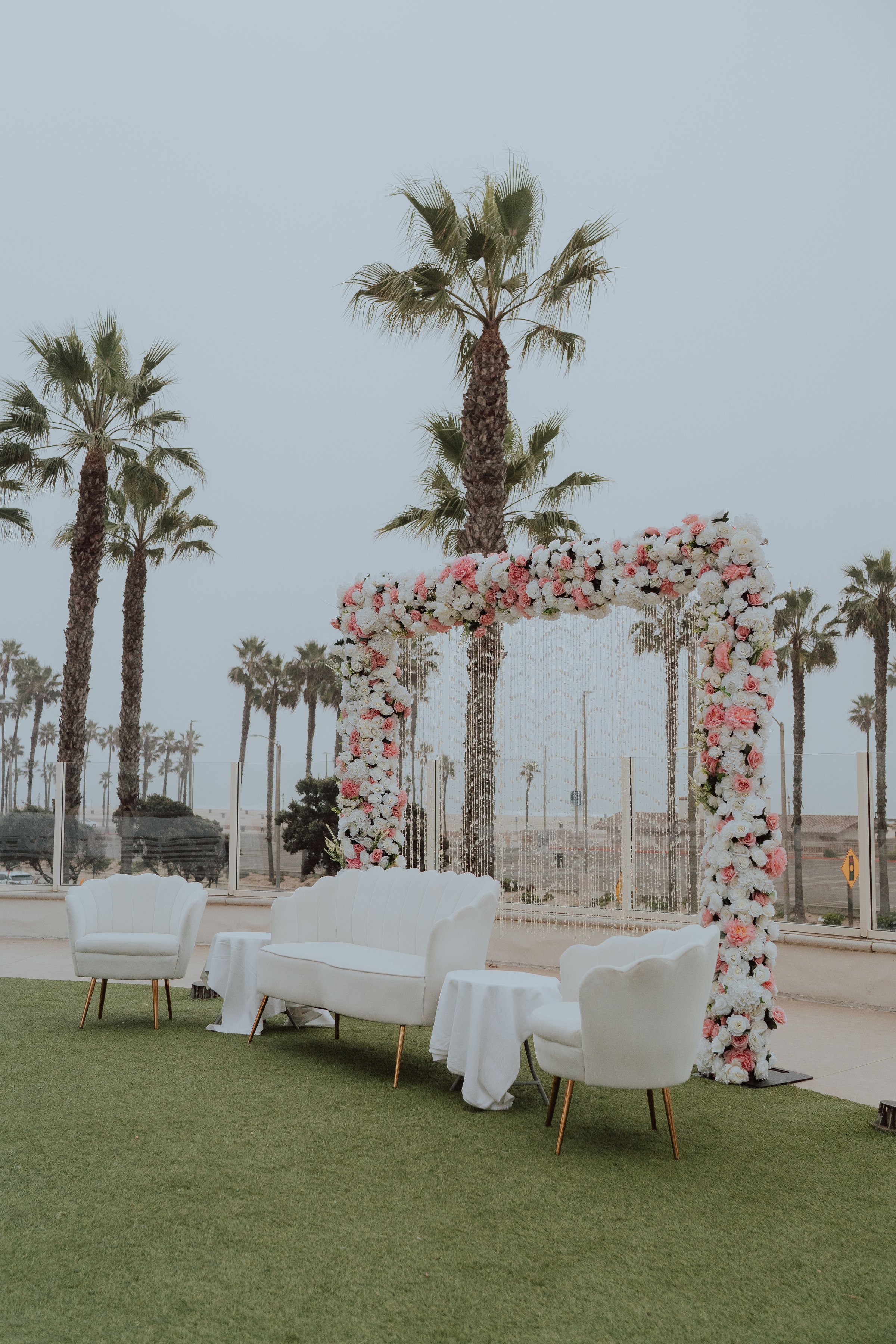 Elegant outdoor wedding setup with white furniture and a floral archway, set against palm trees and a beach background.