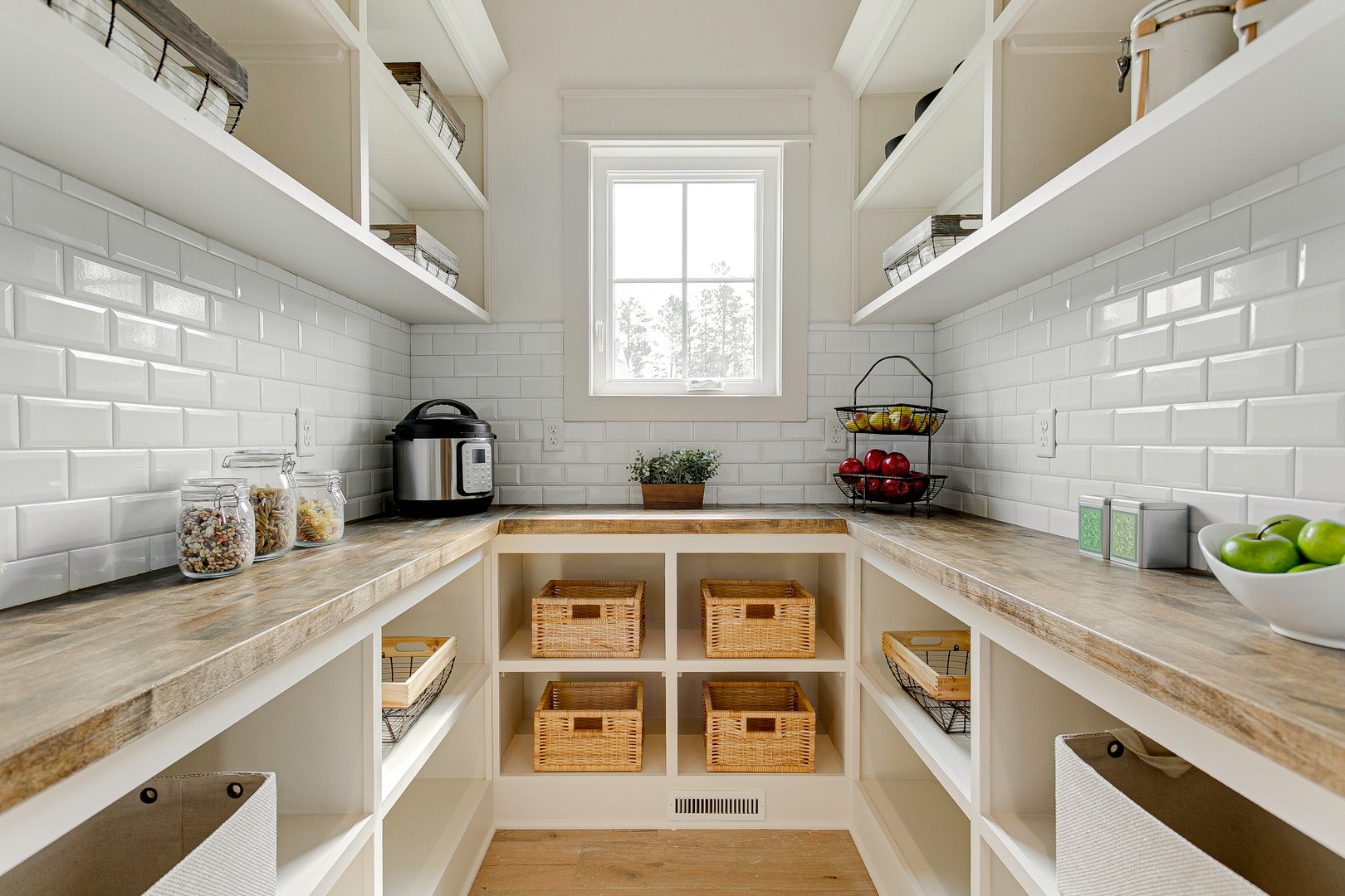 An organized pantry featuring wooden shelves and storage baskets, providing ample space for kitchen essentials. The bright, white subway tile backsplash adds a fresh, clean look, making it easy to find and access items, promoting efficiency and orderliness.