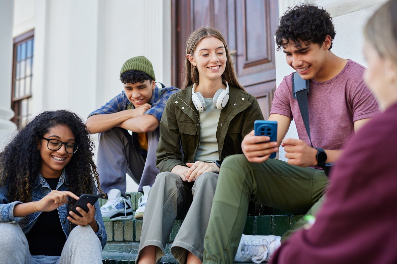 Group of happy students sitting on steps of college campus while looking at mobile phone and smiling. Joyful interaction and bonding captured during a school break outside building. Excited guys and girls laughing and having fun outdoor after lesson while watching video on smartphone.